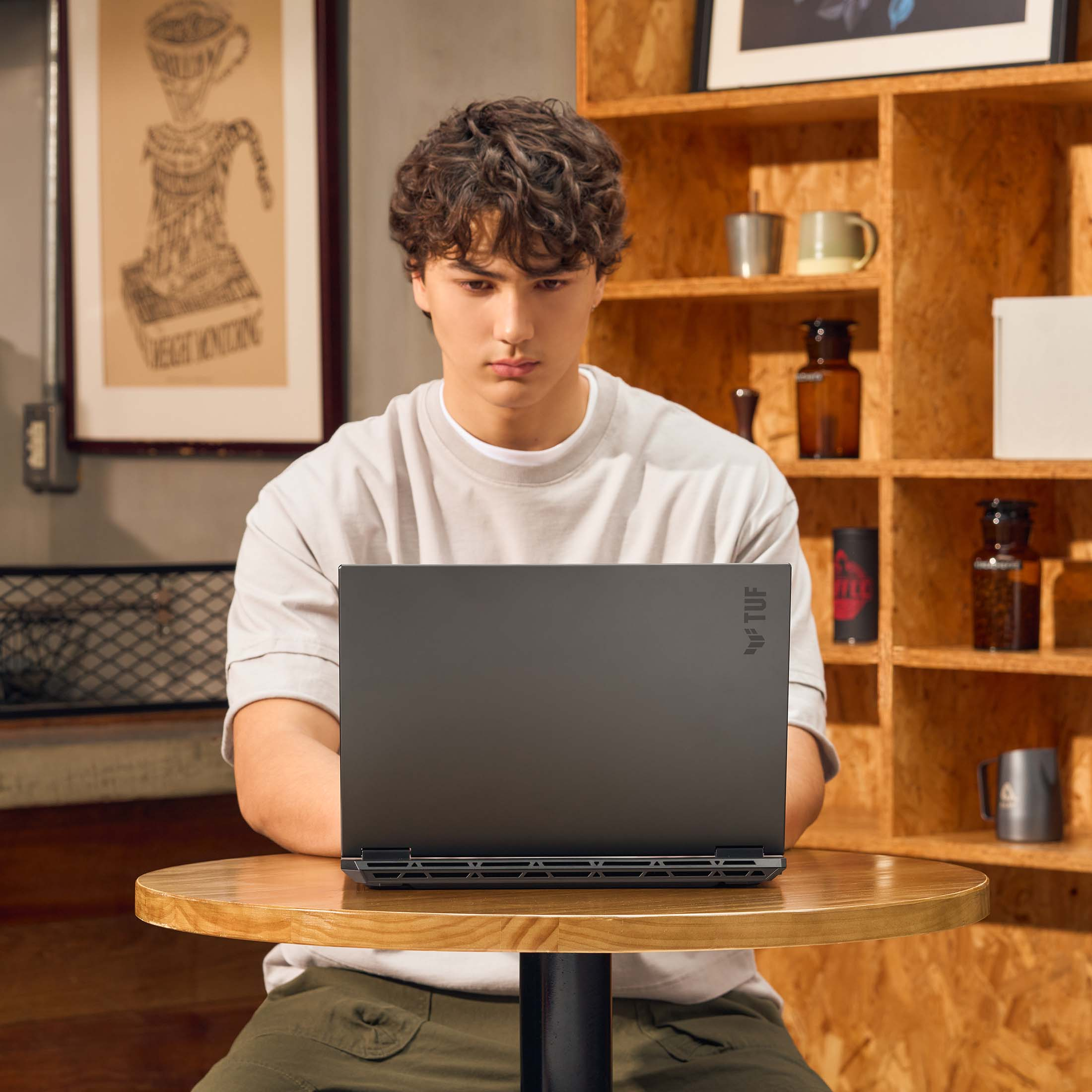 Ein junger Mann sitzt an einem Holztisch und benutzt ein schwarzes Laptop. Im Hintergrund sind Regale mit Gläsern.