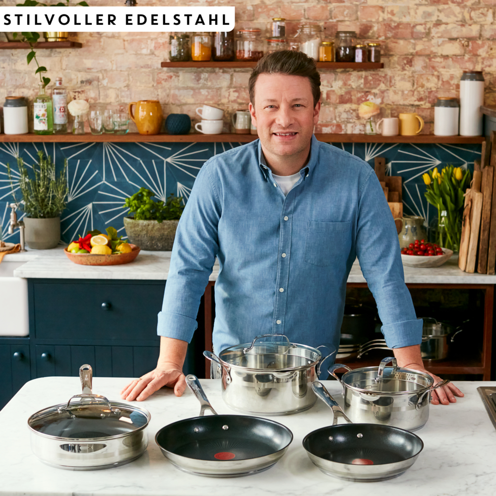 Le chef Jamie Oliver se tient devant un ensemble d'ustensiles de cuisine en acier inoxydable.