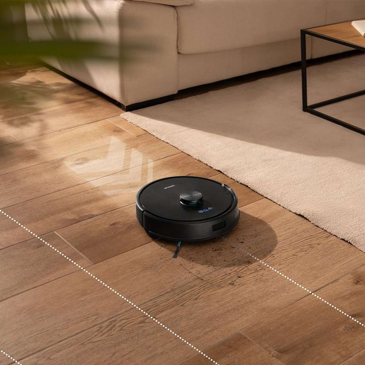 Un robot aspirapolvere nero pulisce un pavimento in legno vicino a un divano e tappeto beige.