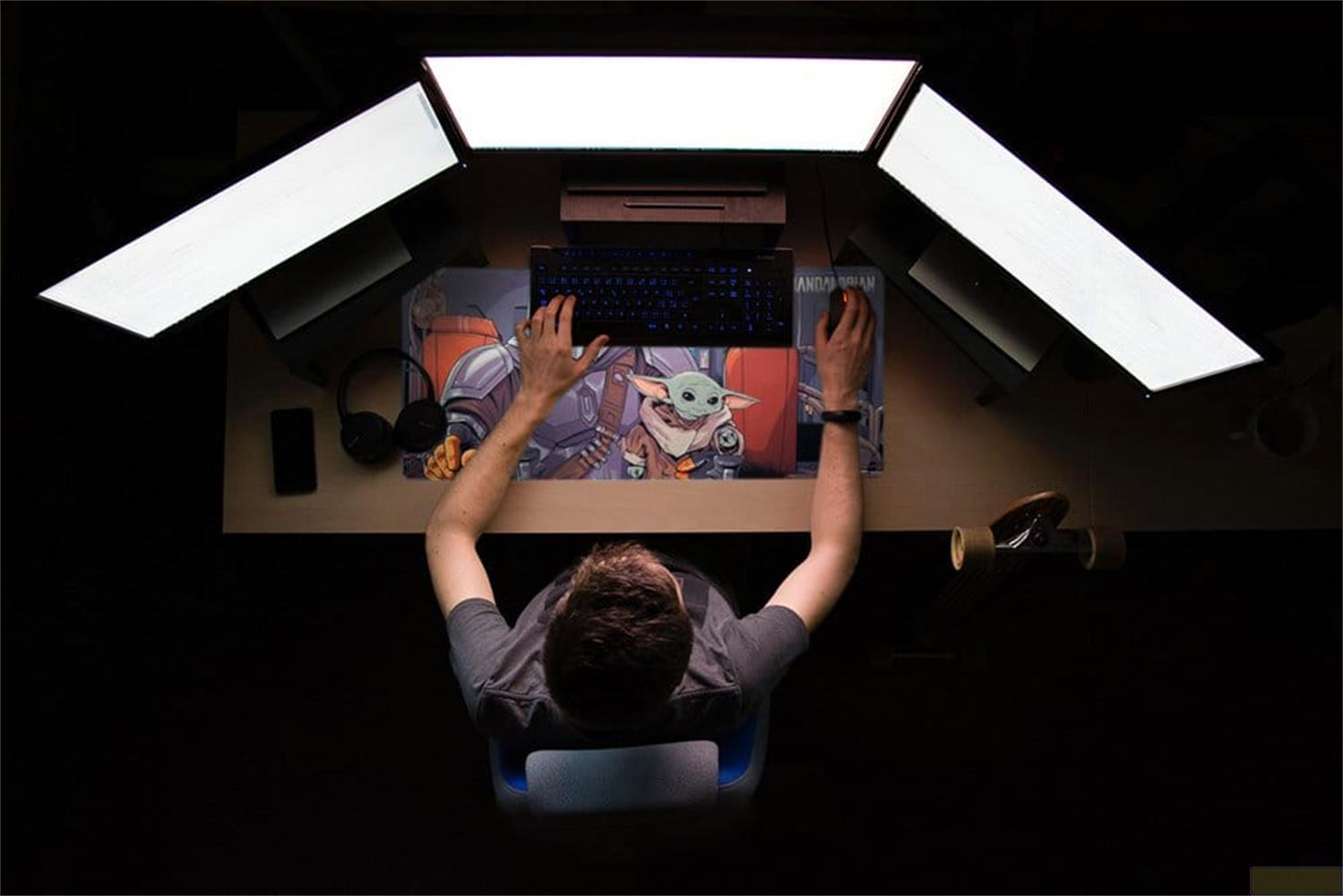 Mężczyzna siedzi przy biurku, grając na trzech monitorach, klawiaturze i myszy.