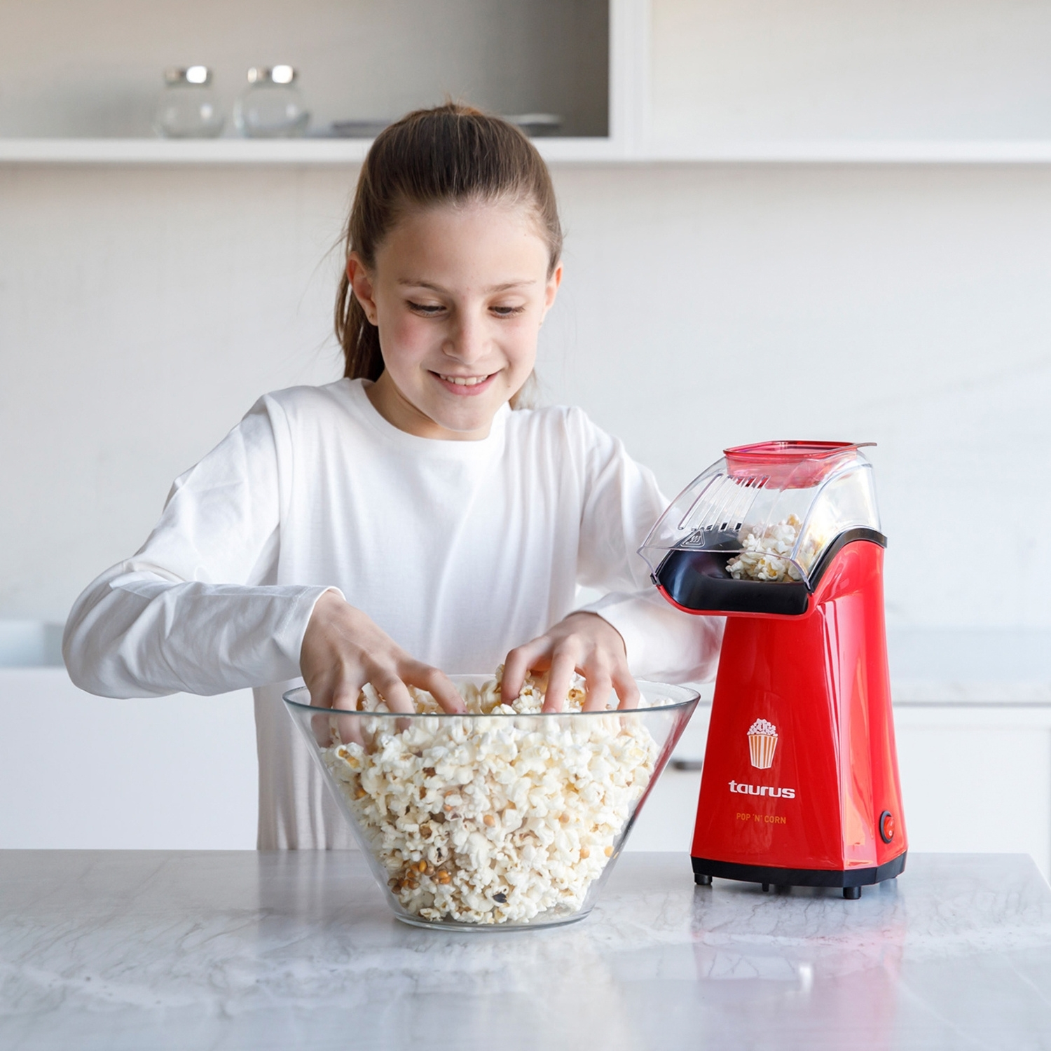 Una niña con un suéter blanco saca palomitas de un tazón junto a una máquina de palomitas roja.