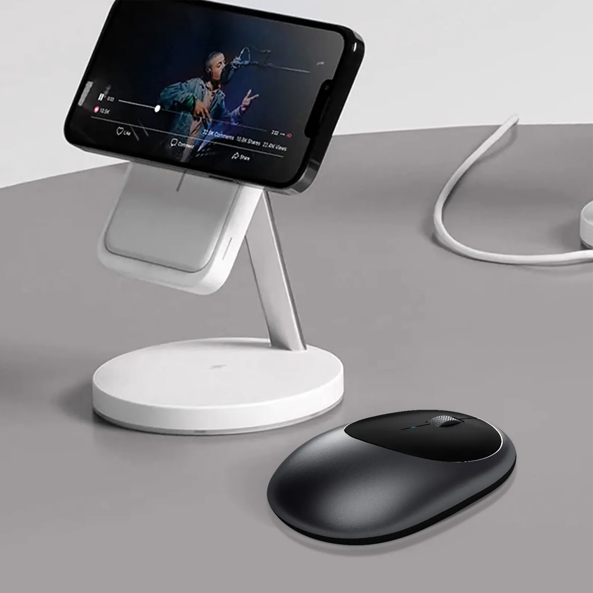 Czarna mysz komputerowa leży na szarym biurku obok telefonu na białej podstawce ładującej.