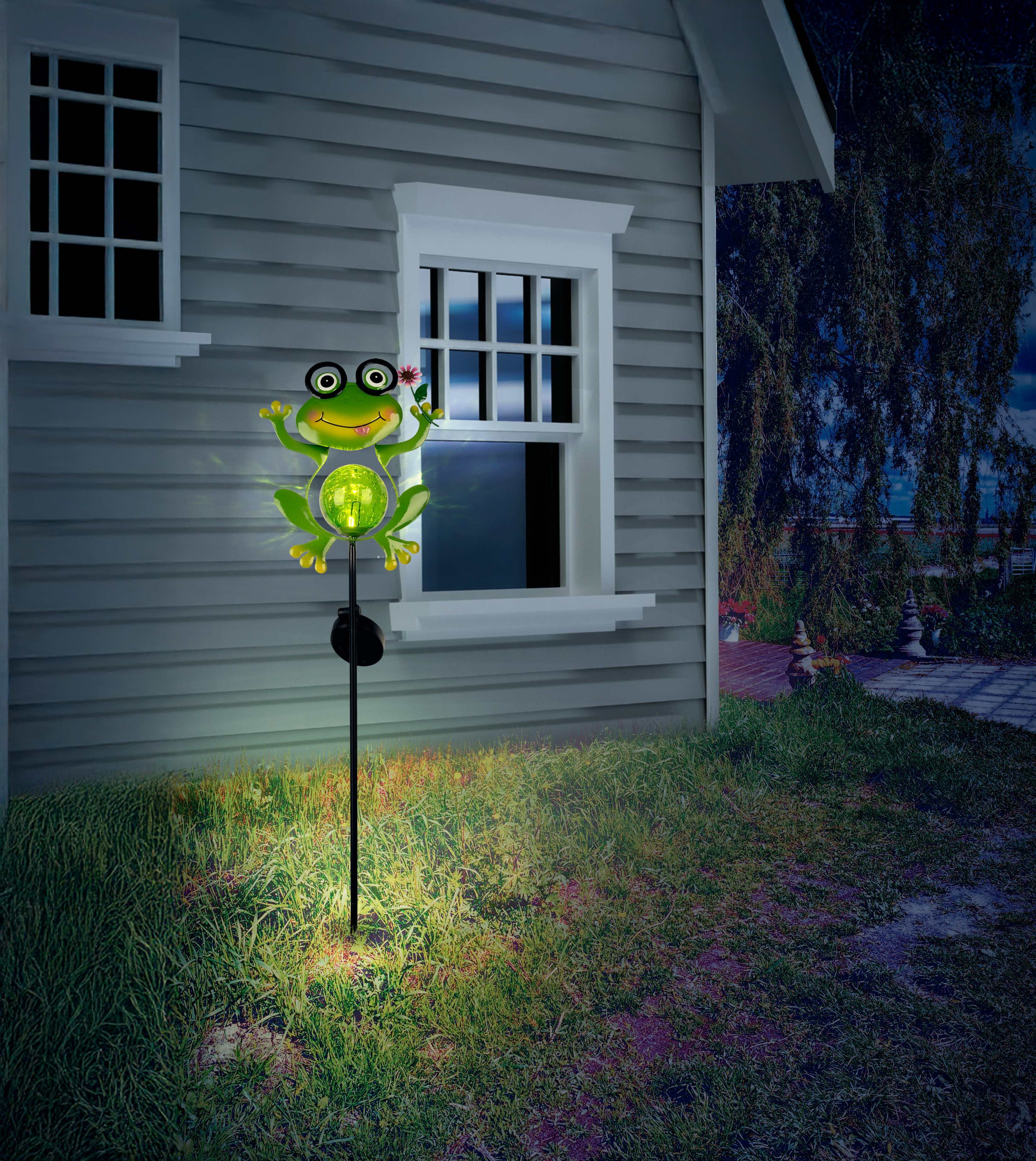 Ein solarbetriebener Frosch mit einer Lampe leuchtet im Gras vor einem Haus.