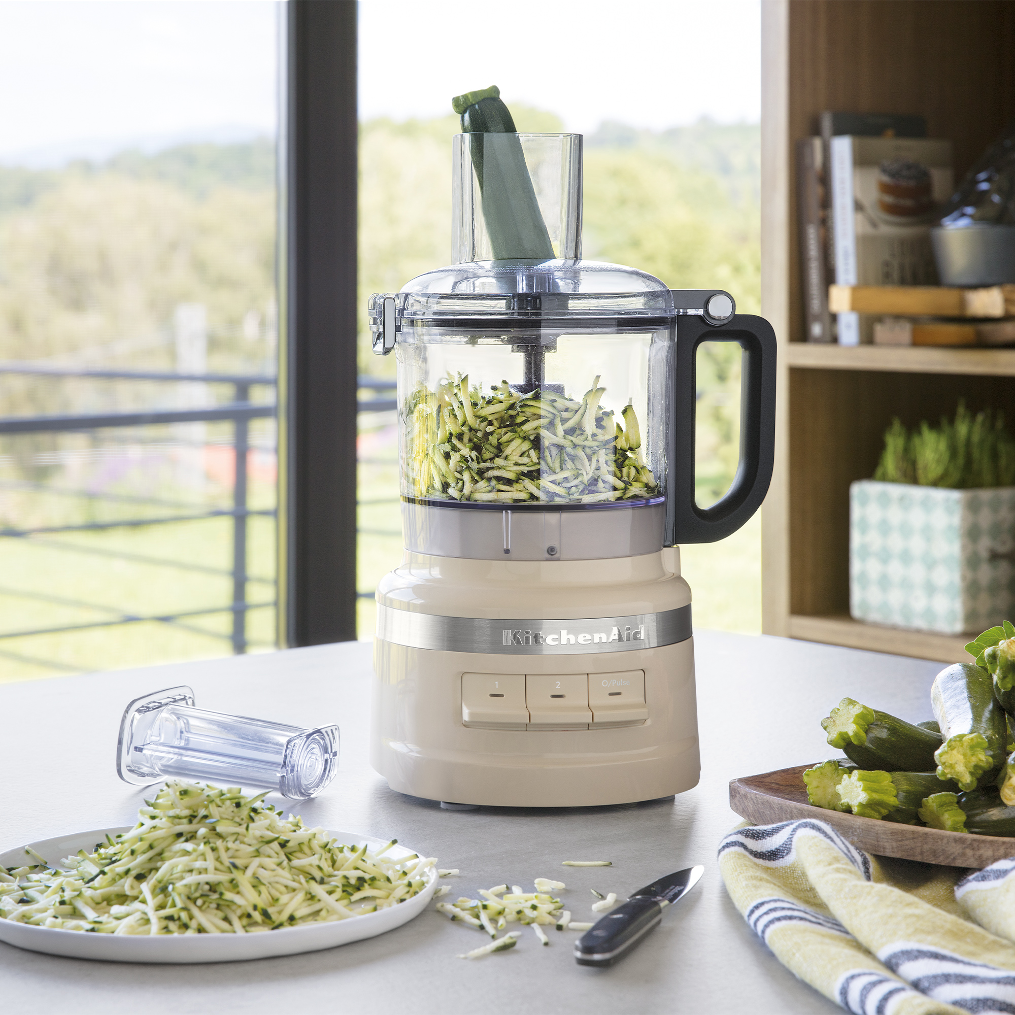 Eine beige Küchenmaschine raspelt Zucchini. Daneben liegen Zucchini und ein Messer.