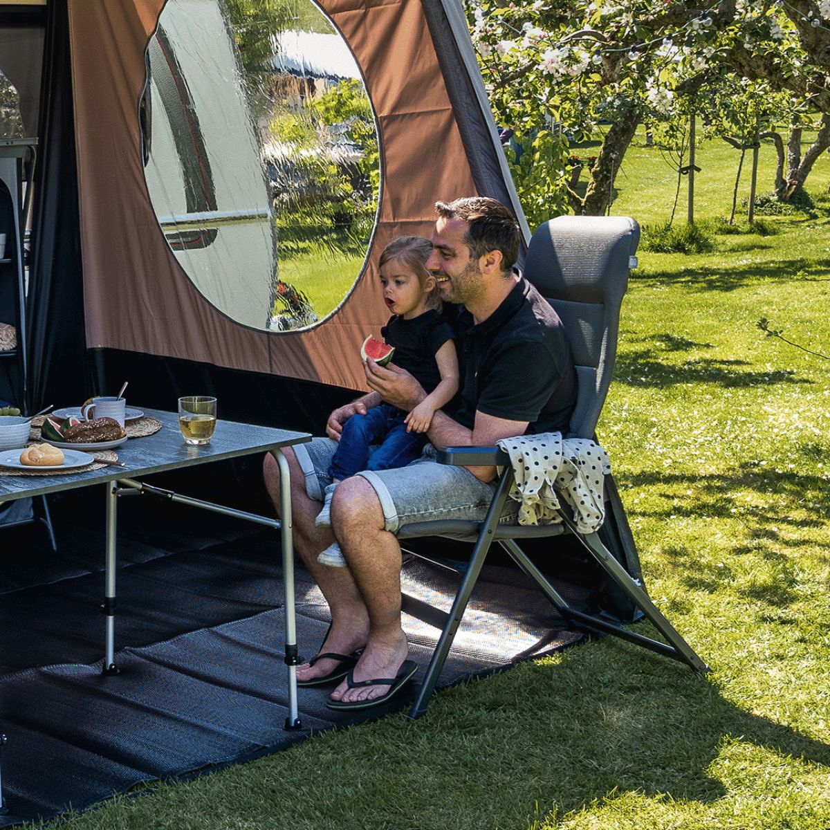 Man met kind eet buiten watermeloen naast een campingtafel met eten.
