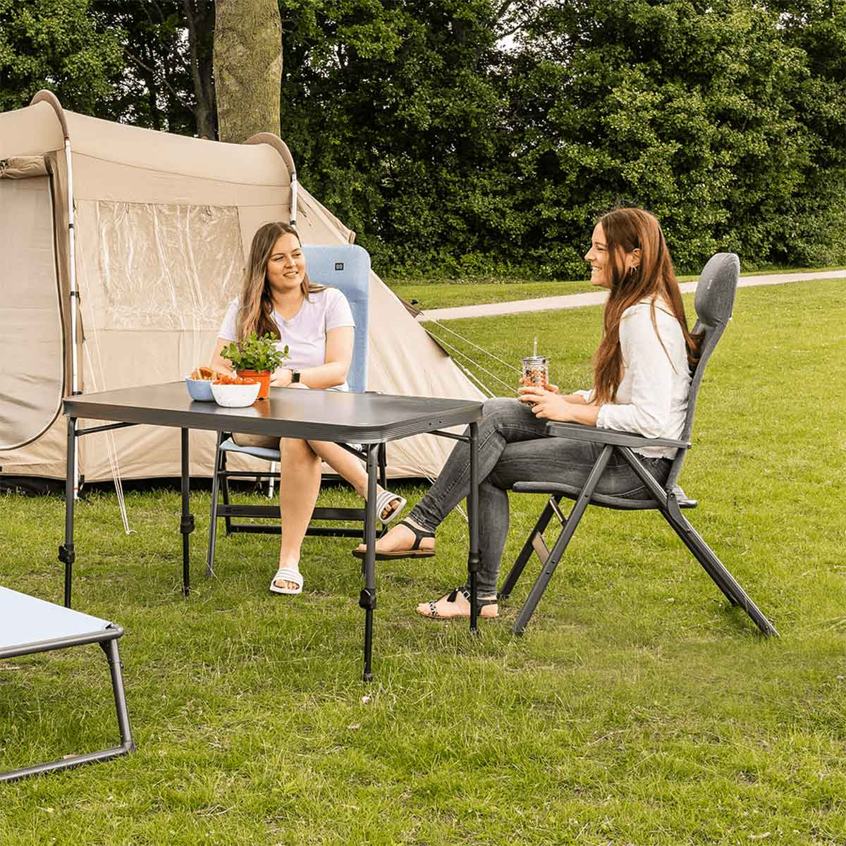 Twee vrouwen zitten buiten aan een tafel naast een tent, op een groen gazon.