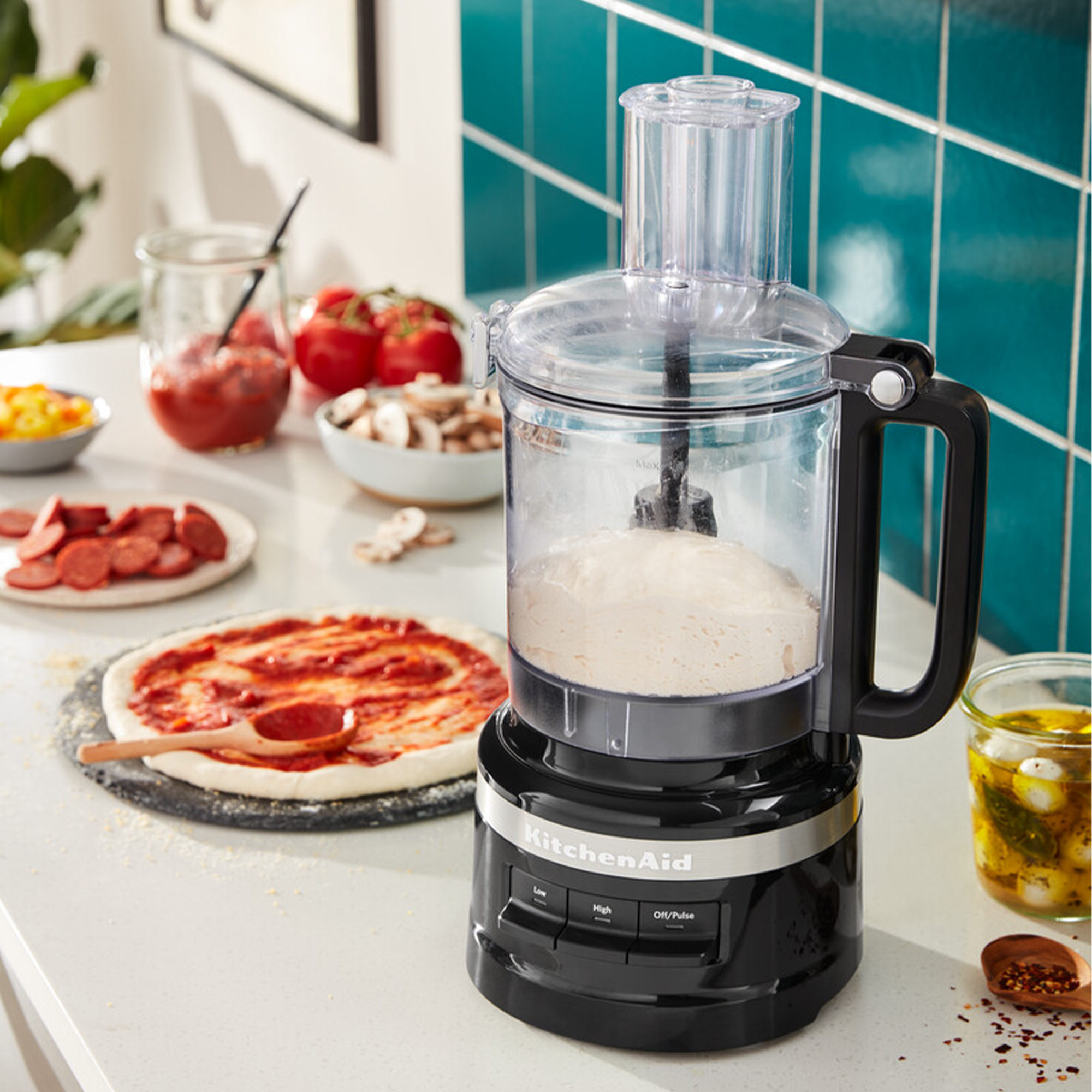Ein Küchenroboter mit Teig steht neben Pizza und Zutaten.