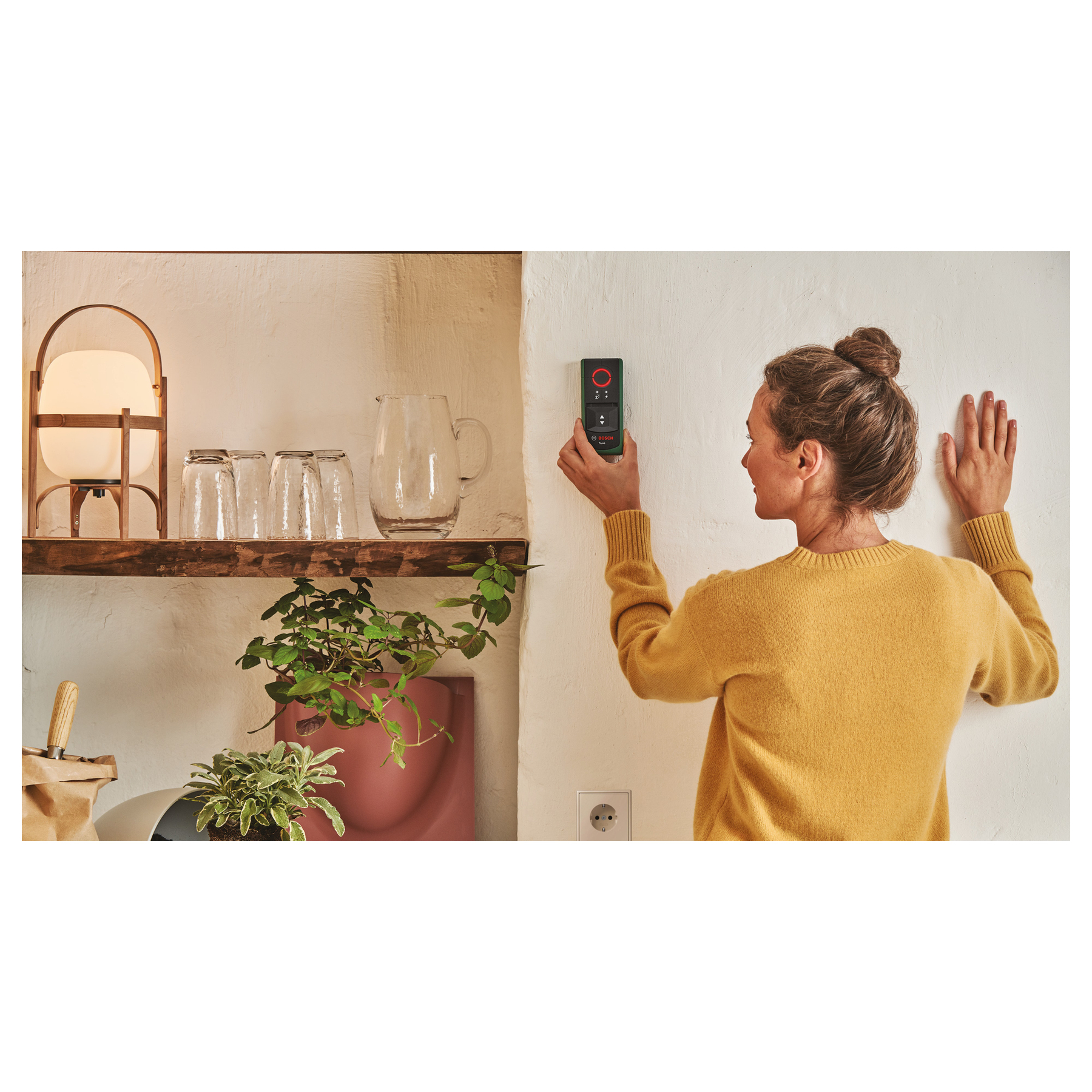 Frau mit gelbem Pullover hält ein Messgerät an eine weiße Wand. Ein Regal mit Gläsern und Pflanzen.