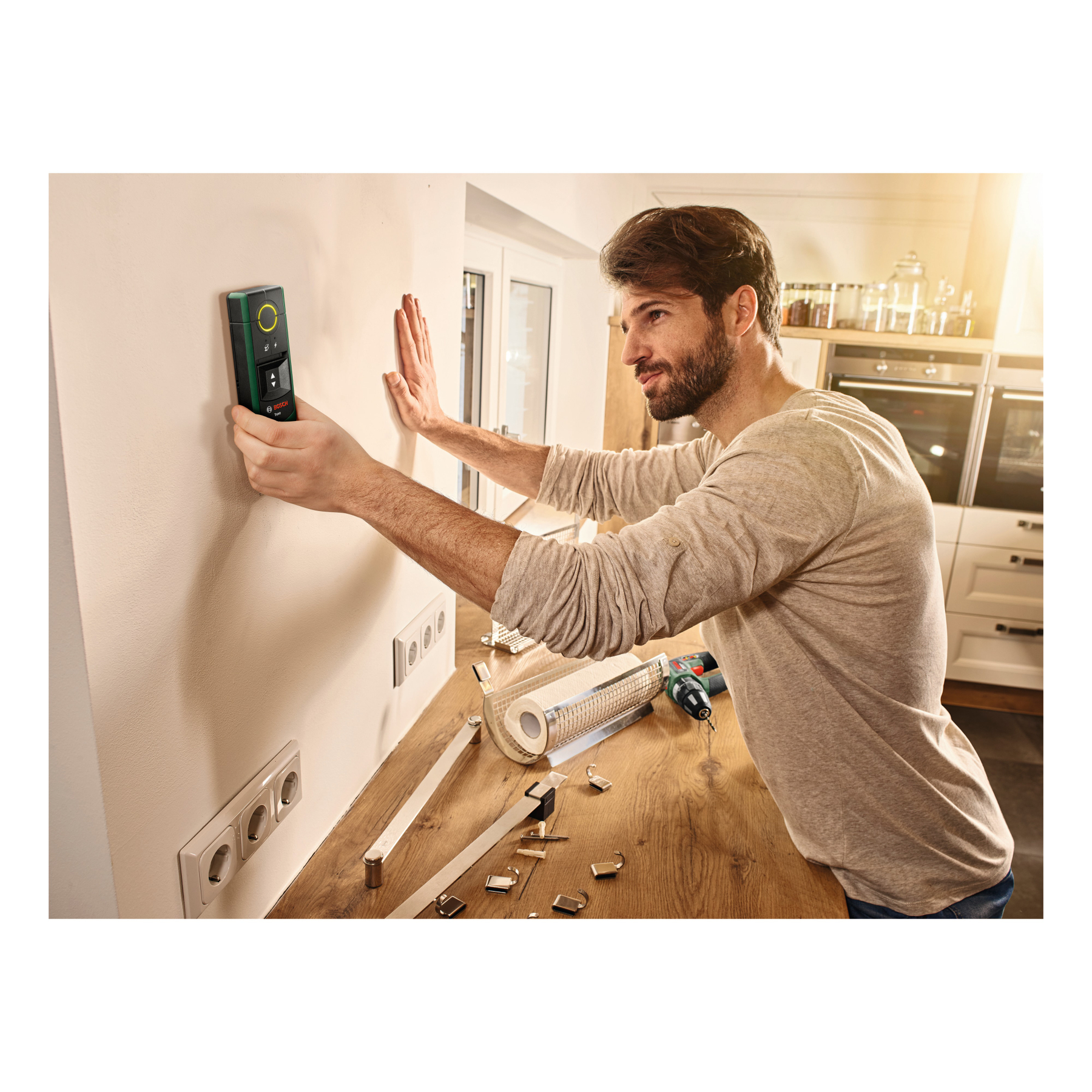 Mann benutzt Messgerät an Wand. Werkzeuge und Küchengeräte im Hintergrund.