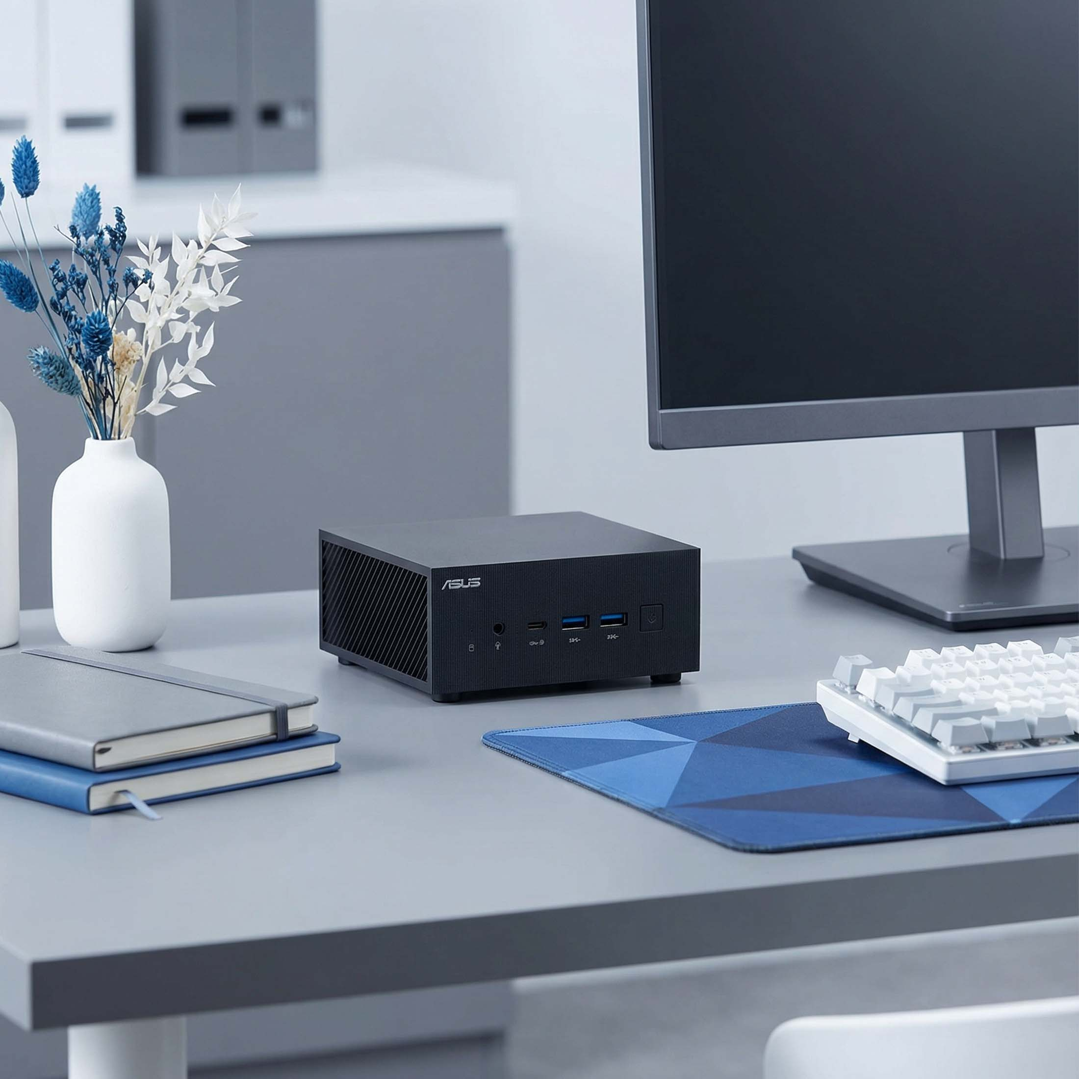 Schwarzer ASUS Mini-PC mit Tastatur, Mauspad, Monitor und Deko auf einem Schreibtisch.