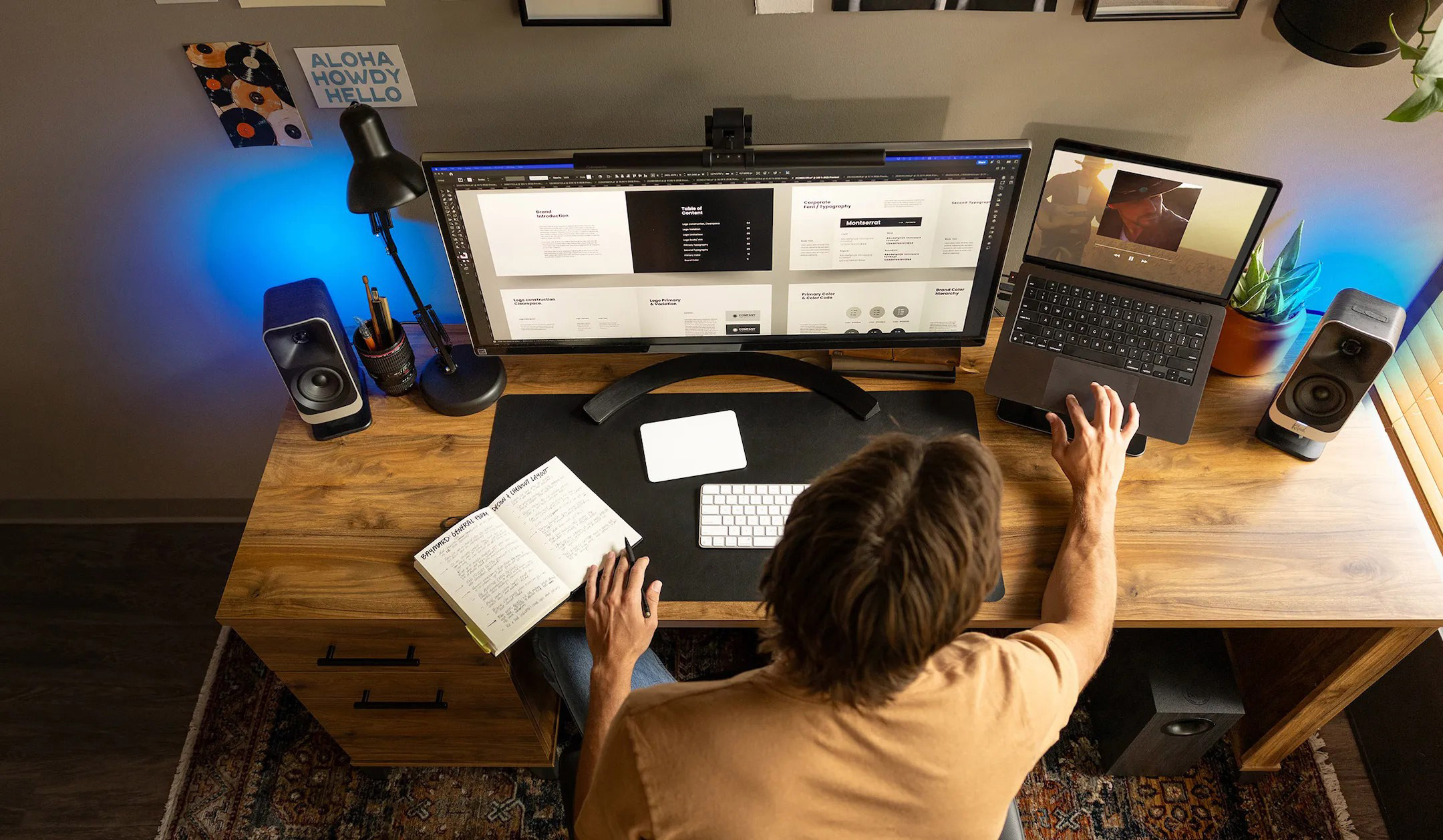 Person am Schreibtisch mit zwei Monitoren, Laptop und Lautsprechern.
