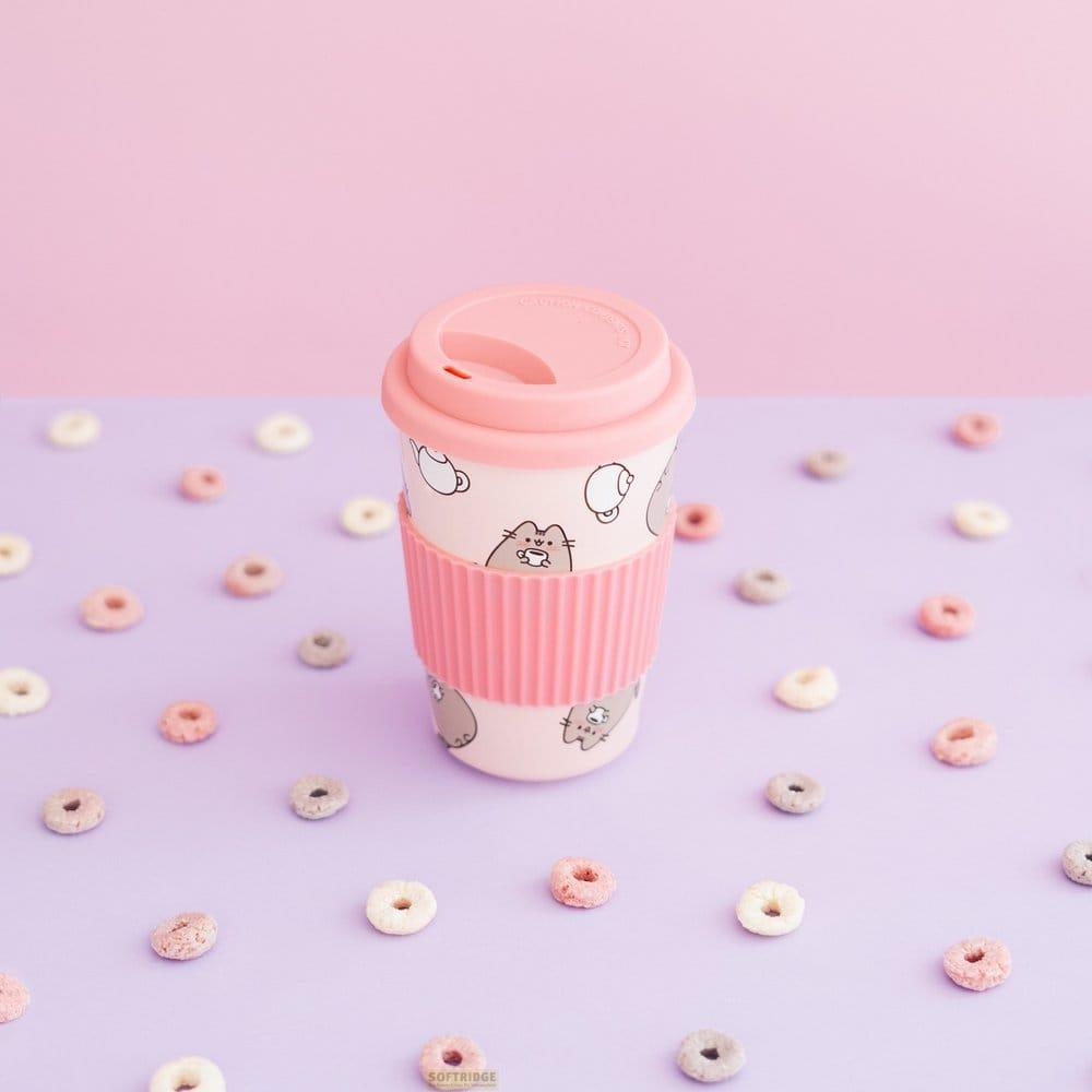 Ein pinker Reisebecher mit Katzenmotiven steht auf einer lilafarbenen Oberfläche, umgeben von bunten Cerealschleifen.