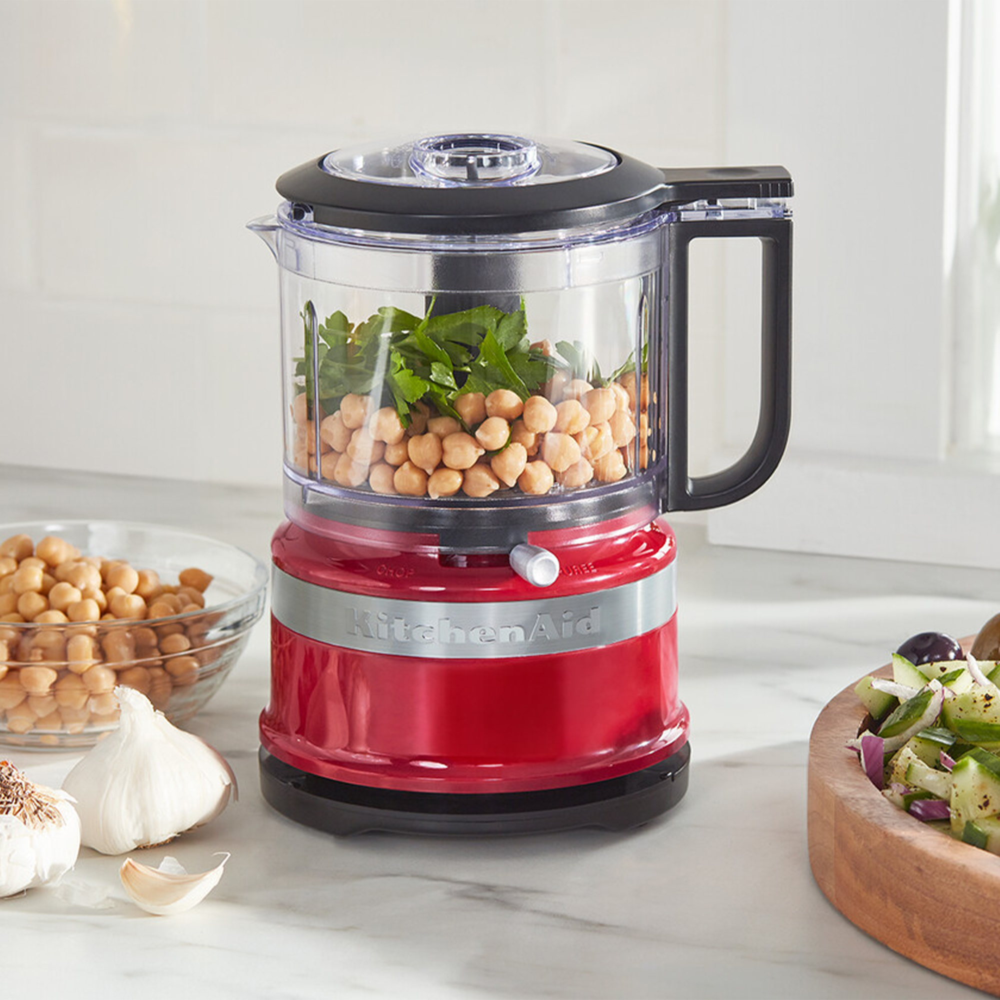 Ein roter KitchenAid-Zerkleinerer mit Kichererbsen und Petersilie, daneben Knoblauch und Salat.