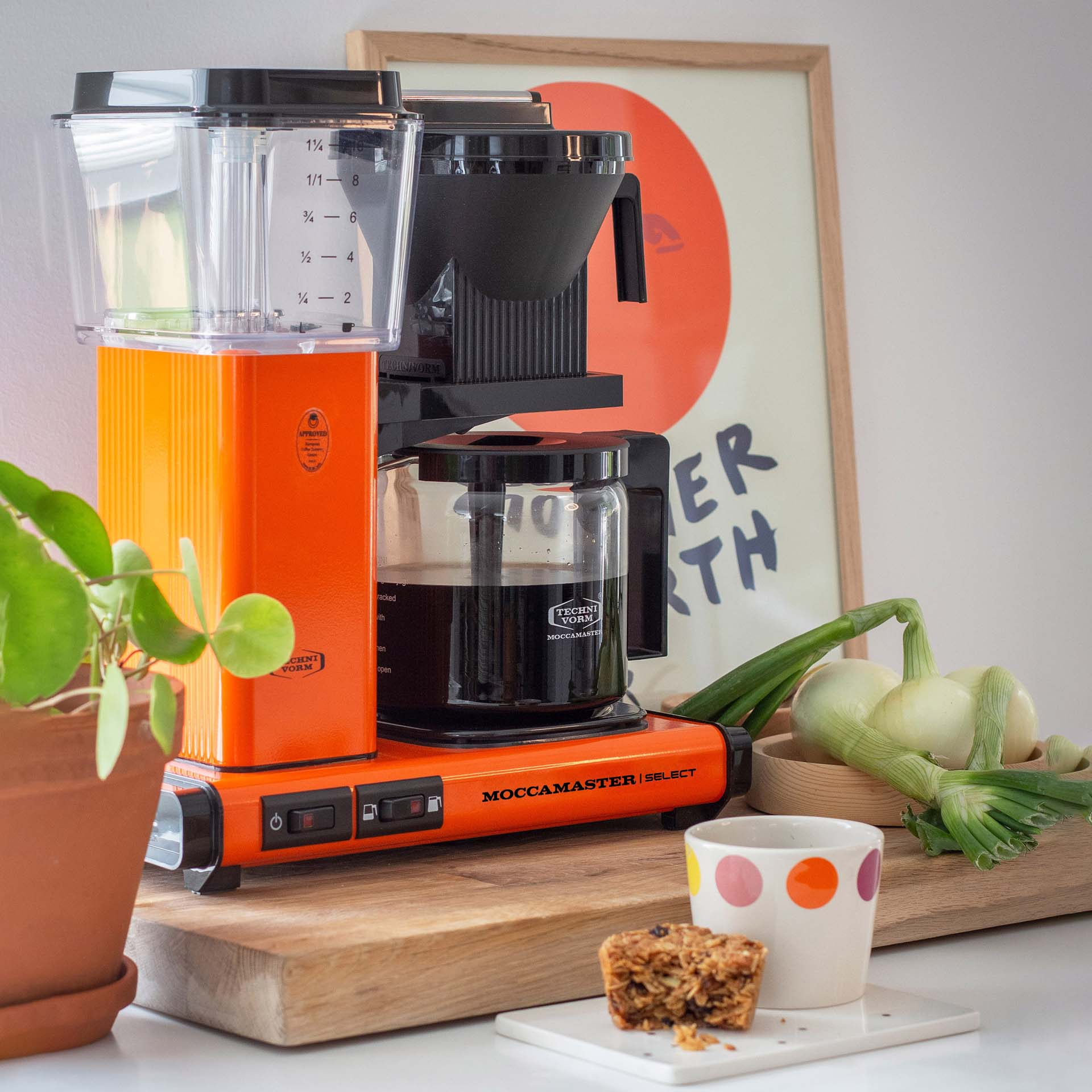 Pomarańczowa ekspres do kawy z dzbankiem, obok cebuli, muffina i rośliny.