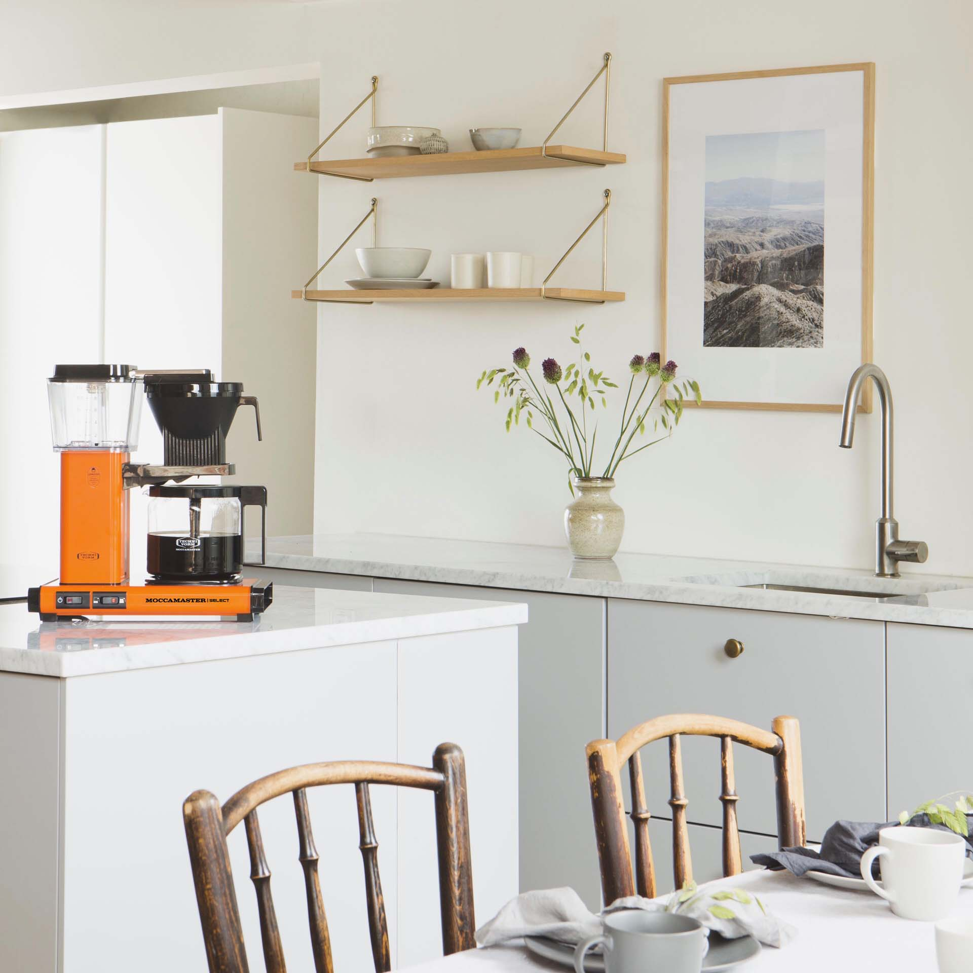 Pomarańczowy ekspres do kawy stoi na marmurowym blacie obok dwóch drewnianych krzeseł.