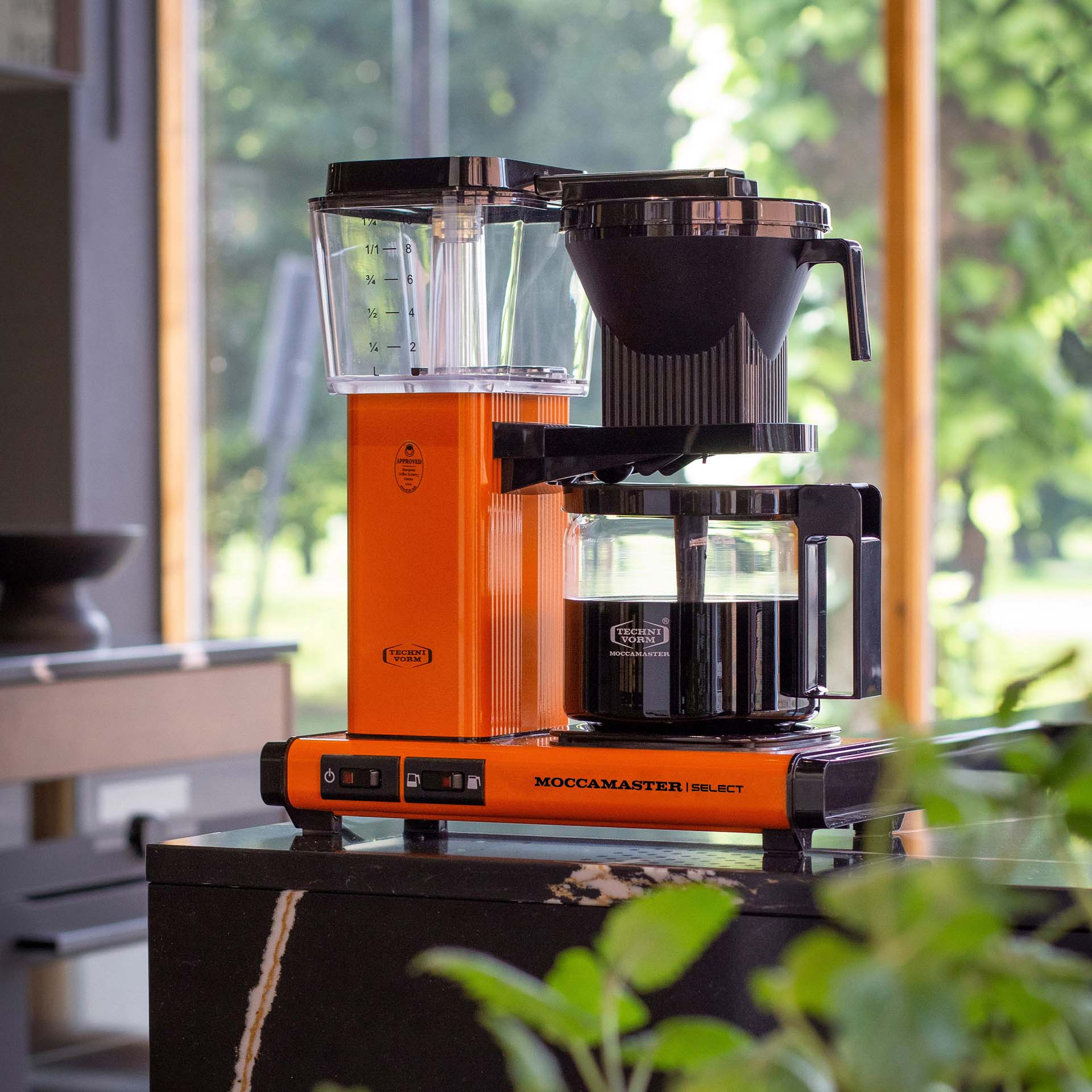 Pomarańczowy ekspres do kawy Moccamaster ze szklanym dzbankiem wypełnionym kawą.