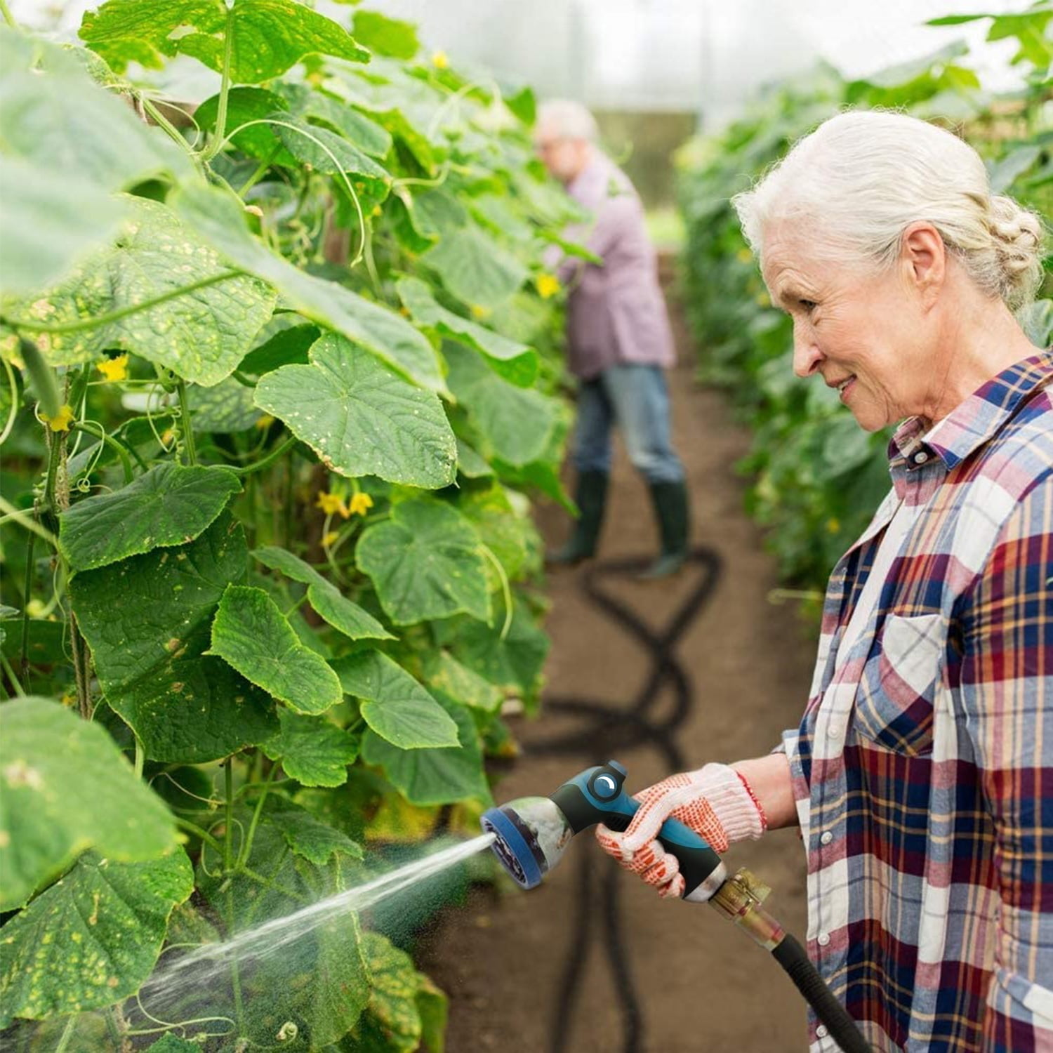 Frau gießt Pflanzen mit Gartenschlauch, Mann im Hintergrund.