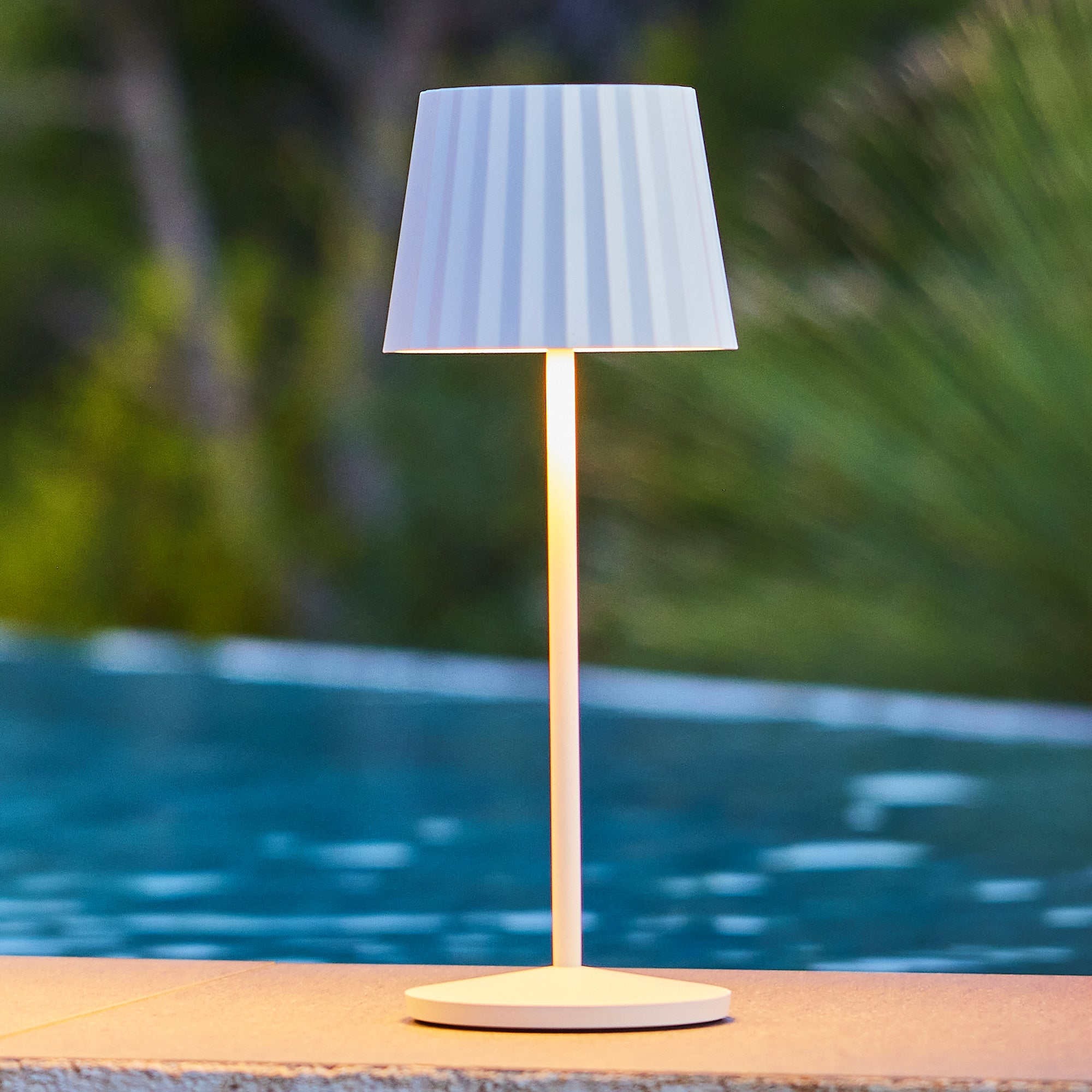 Eine weiße Stehlampe mit geripptem Schirm steht auf einer Steinplatte neben einem Pool.