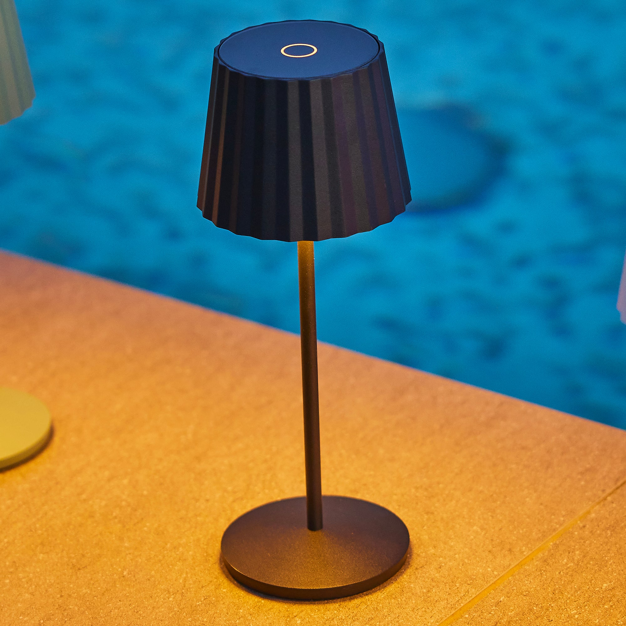 Eine dunkle Lampe mit geripptem Schirm steht auf einem Tisch am Pool.