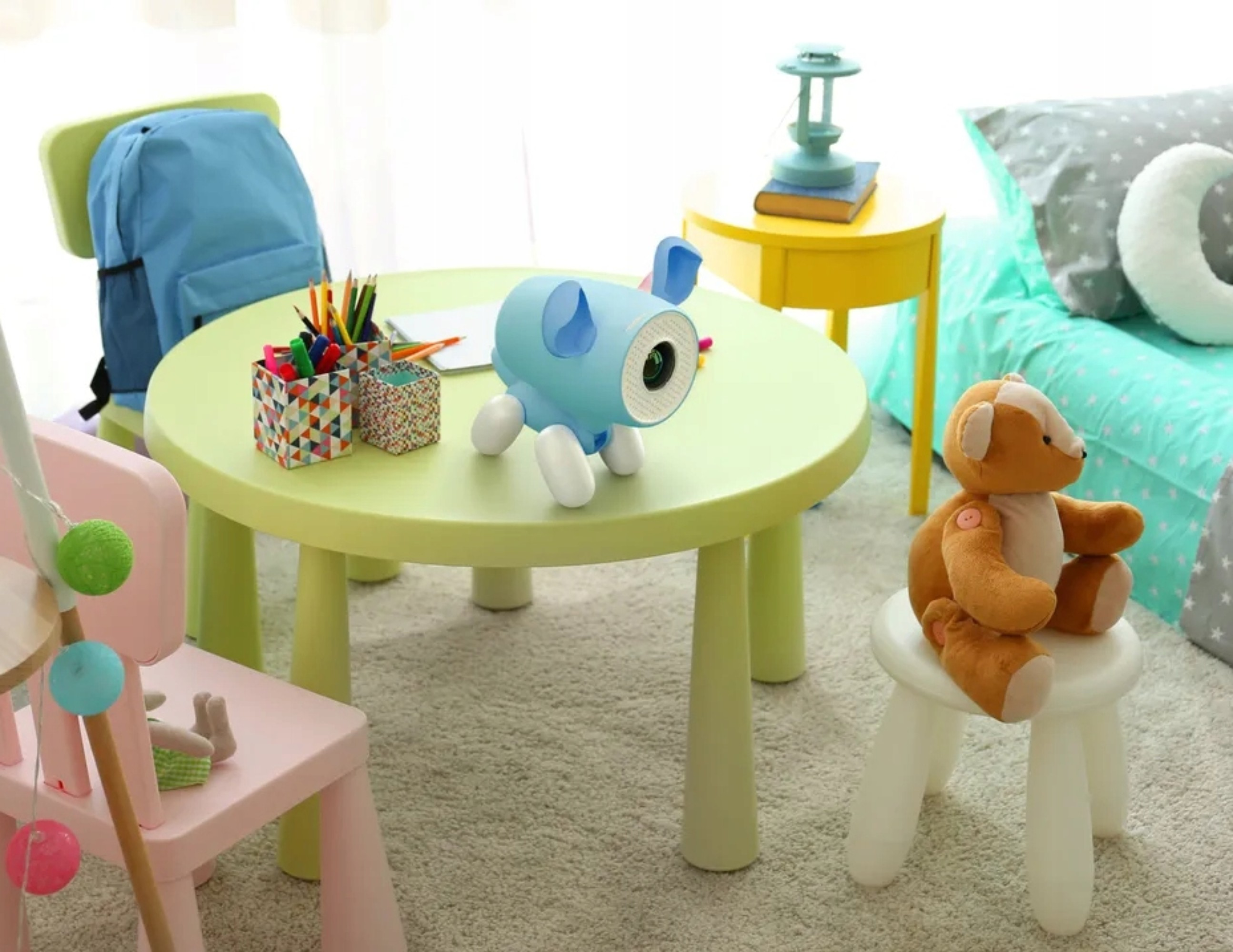 Un juguete de elefante azul sobre una mesa con lápices, sillas y un osito de peluche.