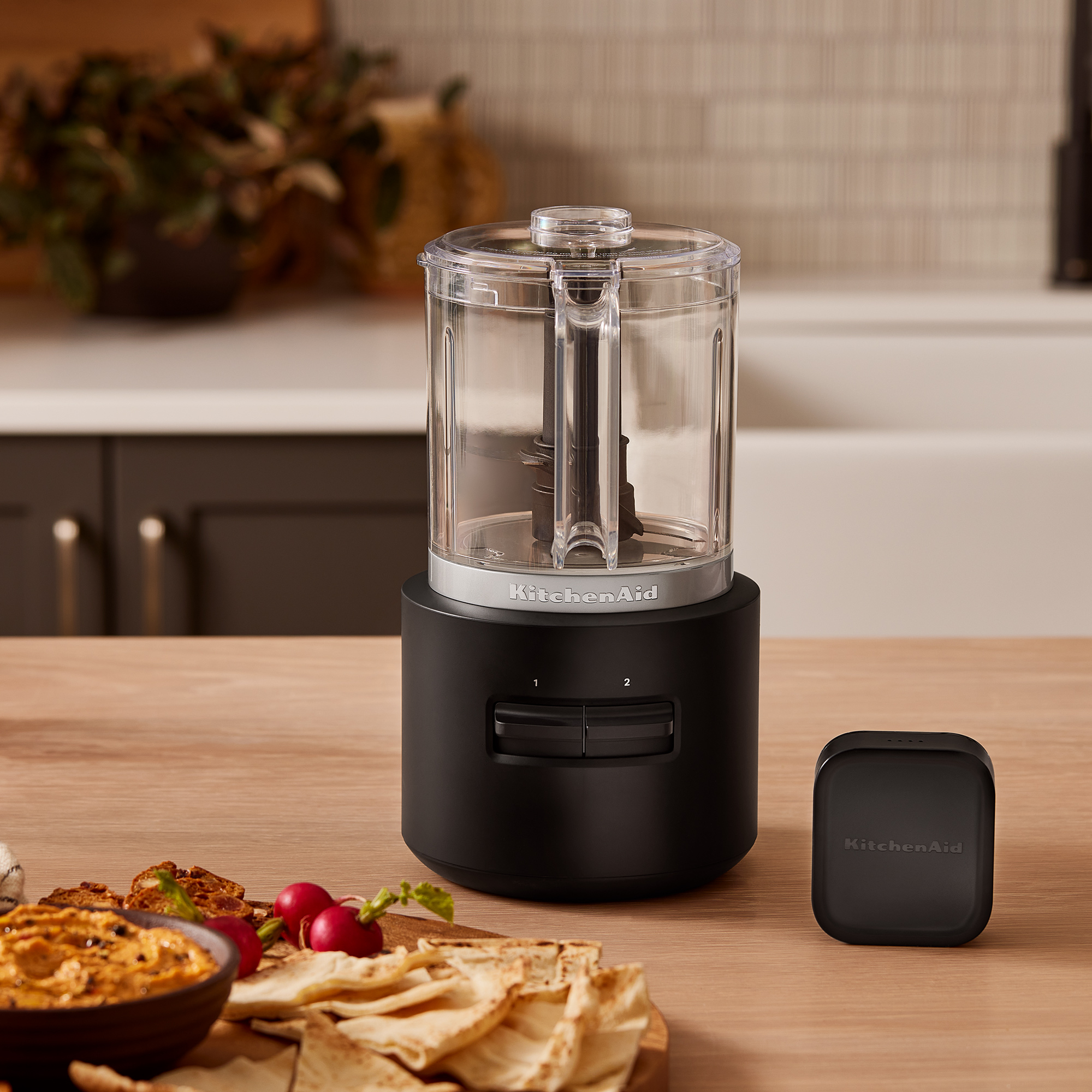 Un mini food processor KitchenAid nero su tavolo di legno con hummus e pane pita.