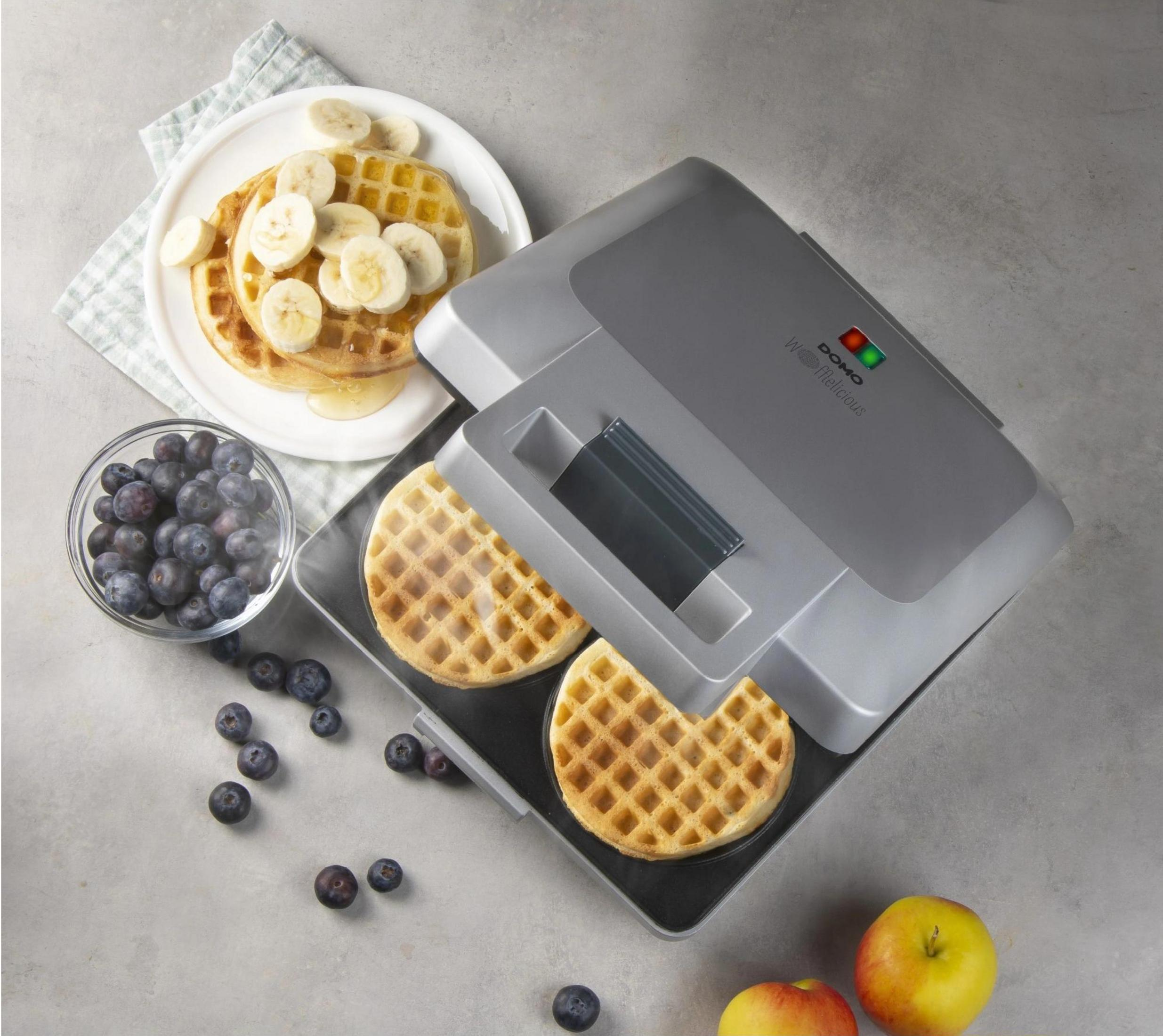 Ein Waffeleisen mit zwei Waffeln, Blaubeeren, Bananenscheiben und Äpfeln.