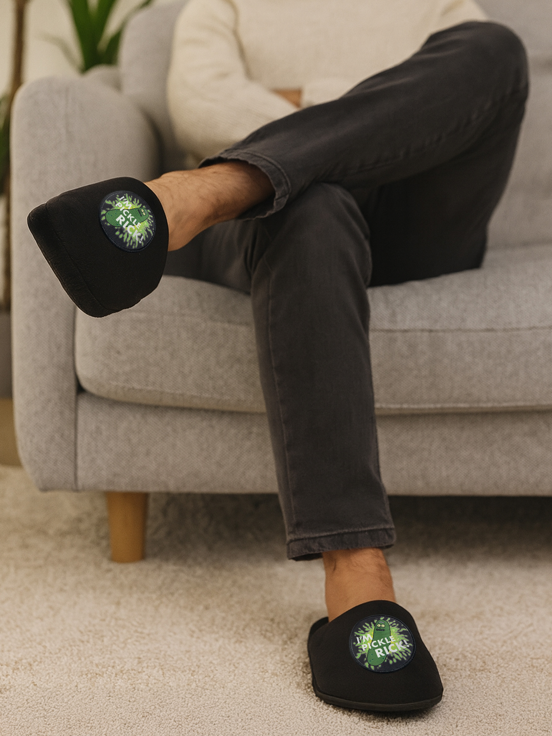 Schwarze Hausschuhe mit Pickle Rick-Aufnäher auf einem Mann, der auf einem Sofa sitzt.