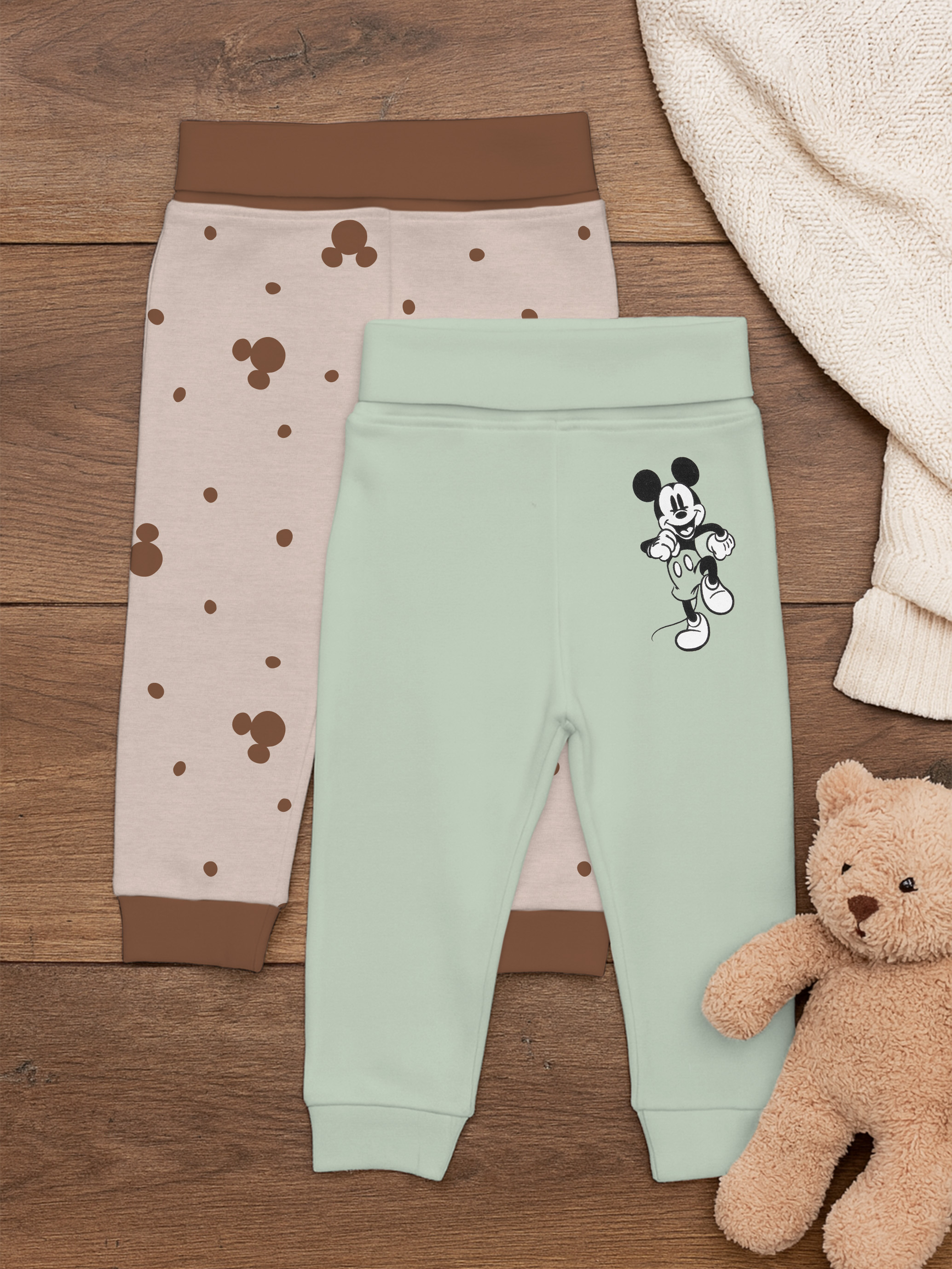 Zwei Babyhosen auf Holzuntergrund, eine mit Micky-Maus, eine mit Punkten.