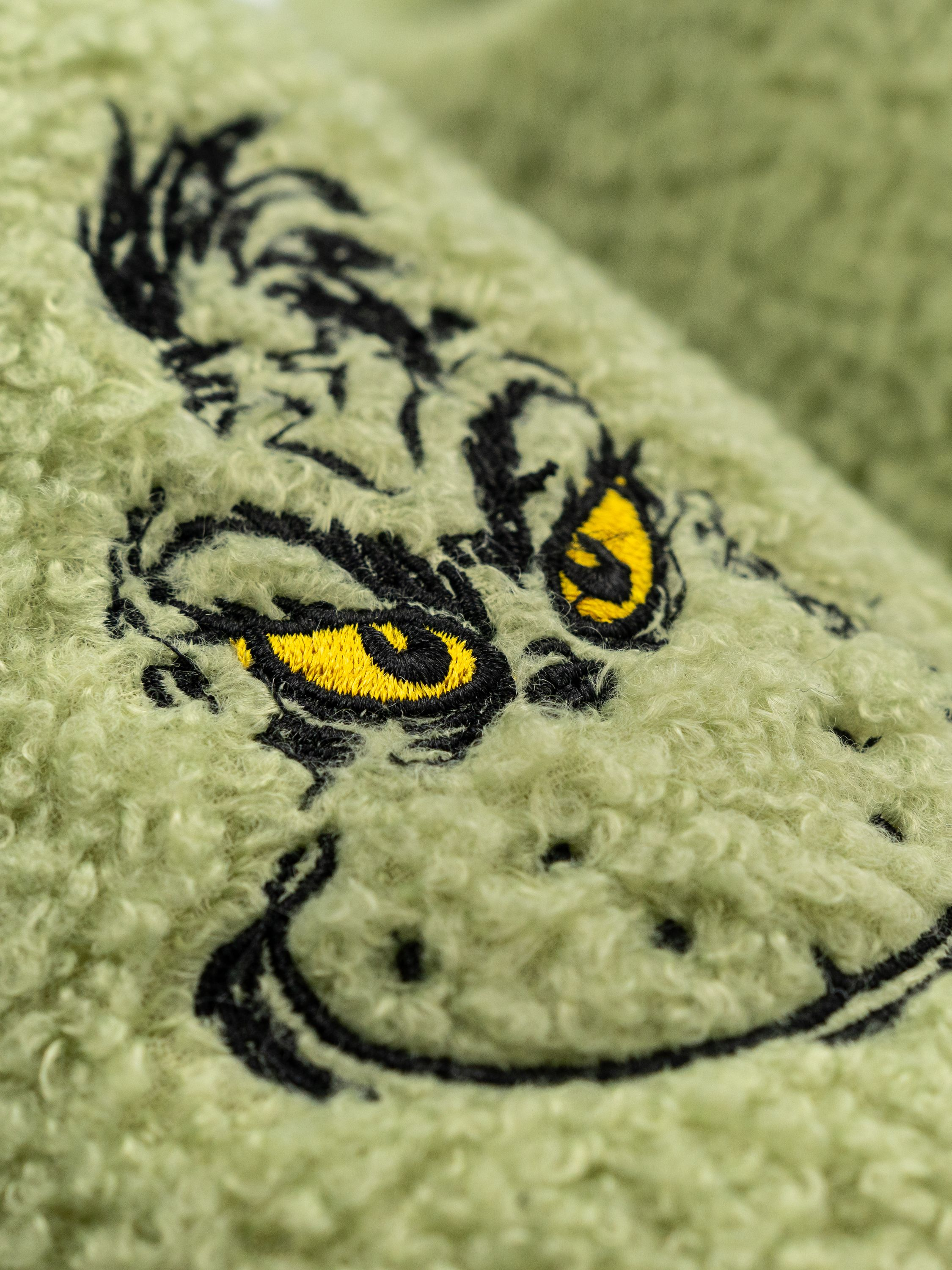 Grinch-Gesicht auf hellgrünem Teddy-Stoff, schwarze Konturen, gelbe Augen.