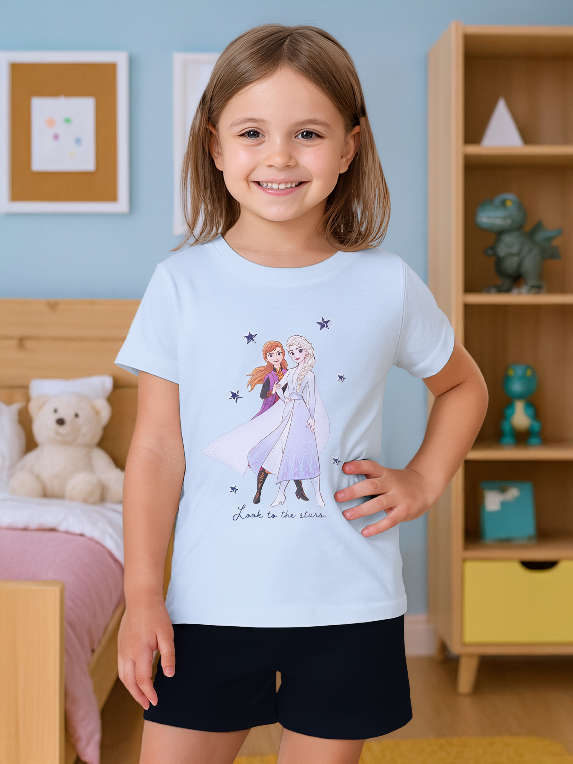 Ein Mädchen in einem hellblauen T-Shirt mit Frozen-Aufdruck steht vor einem Bett.