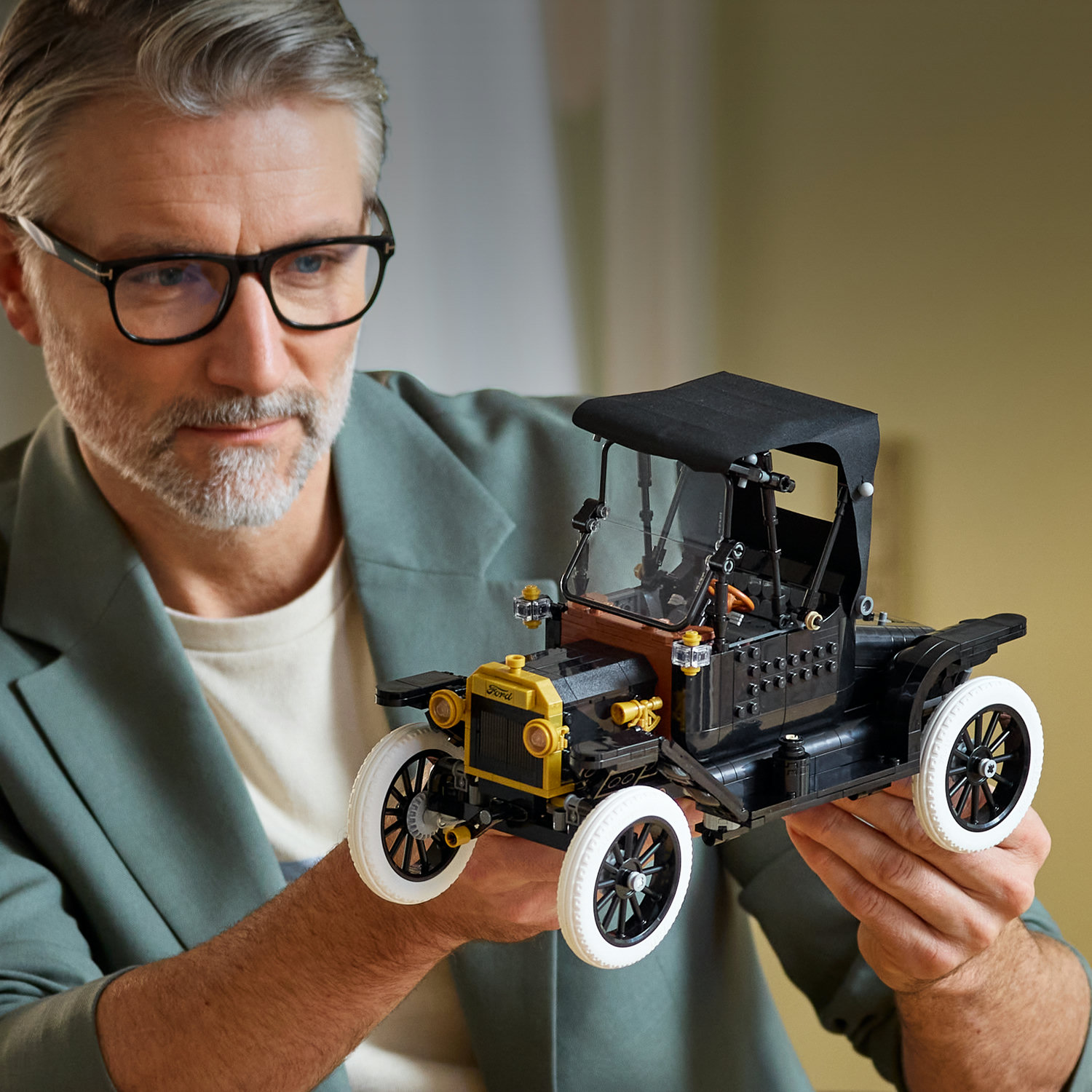 Mężczyzna w okularach trzyma czarny model samochodu Lego z żółtą kratką i białymi oponami.