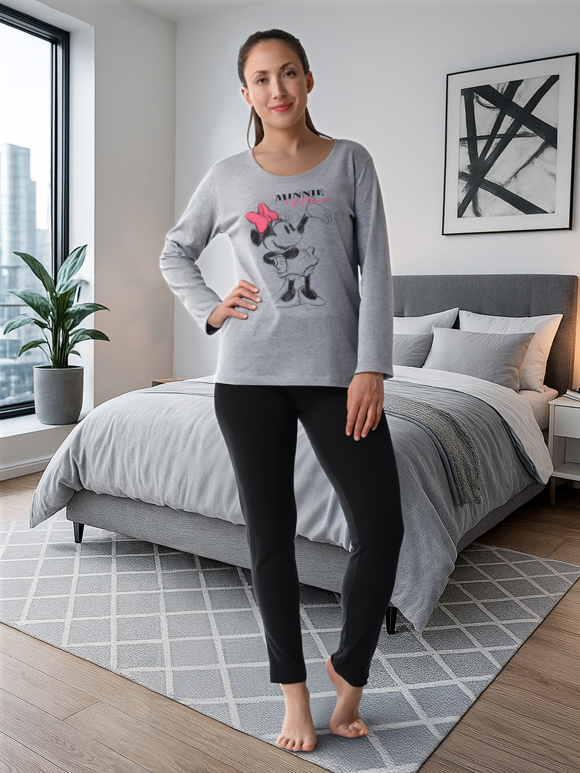 Eine Frau in grauer Minnie Maus Pyjamahose und schwarzer Hose steht vor einem Bett.