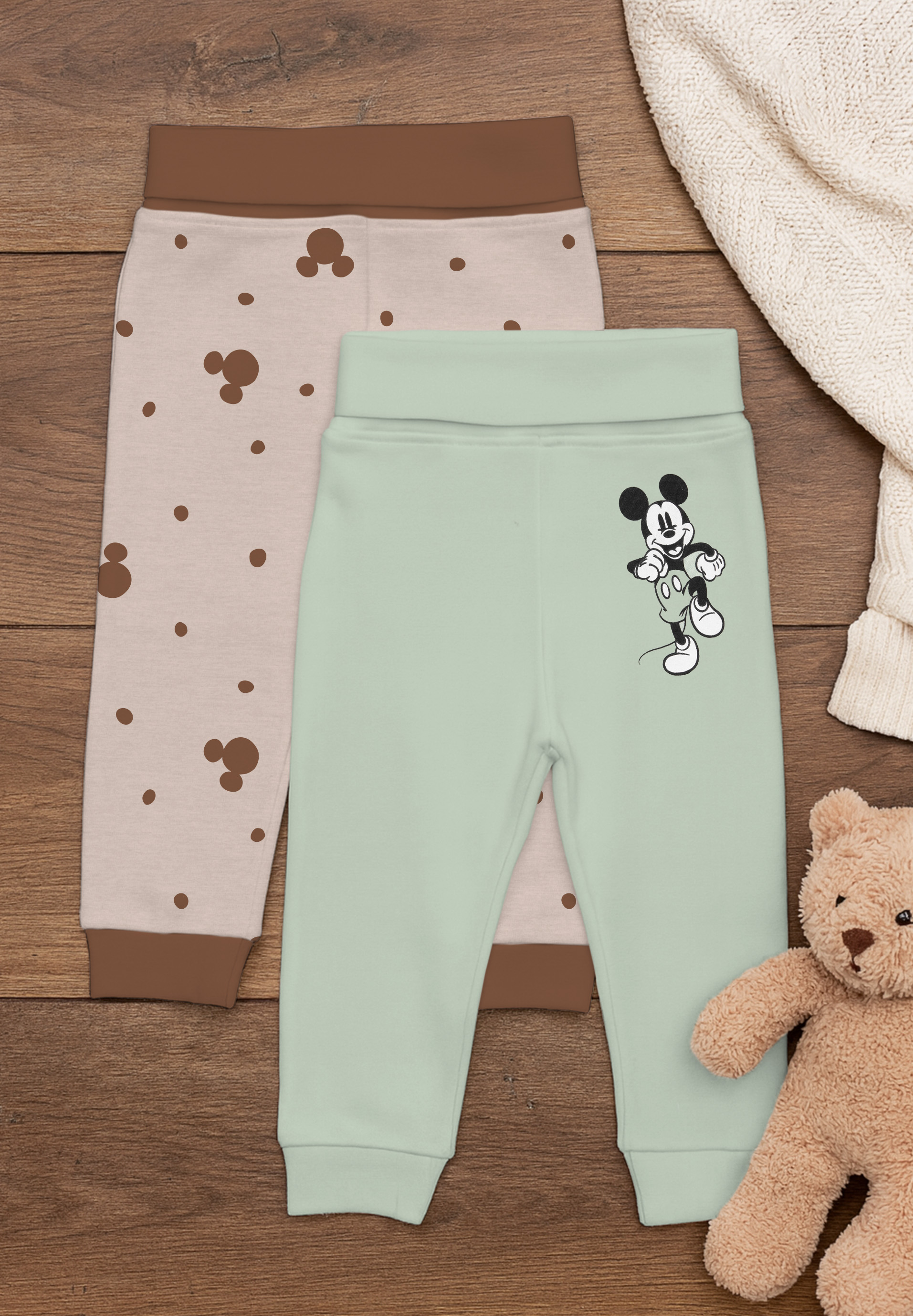 Zwei Babyhosen auf Holzhintergrund, eine hellgrün mit Micky Maus, eine beige mit Punkten.