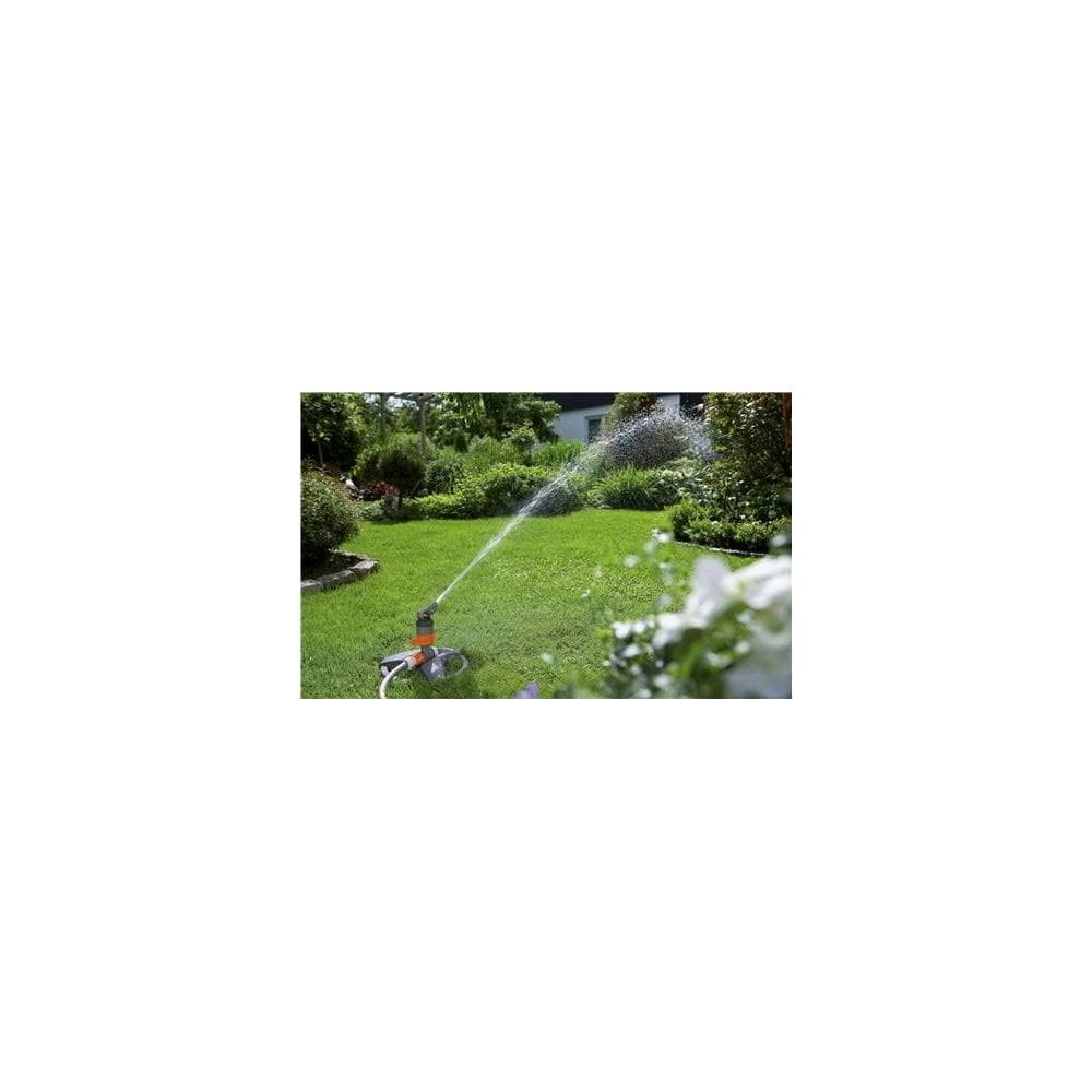 Ein Sprinkler bewässert einen grünen Rasen in einem Garten mit Bäumen und Büschen im Hintergrund.