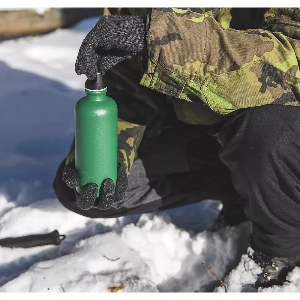 Person in Tarnjacke und Handschuhen hält grüne Flasche in verschneiter Umgebung.