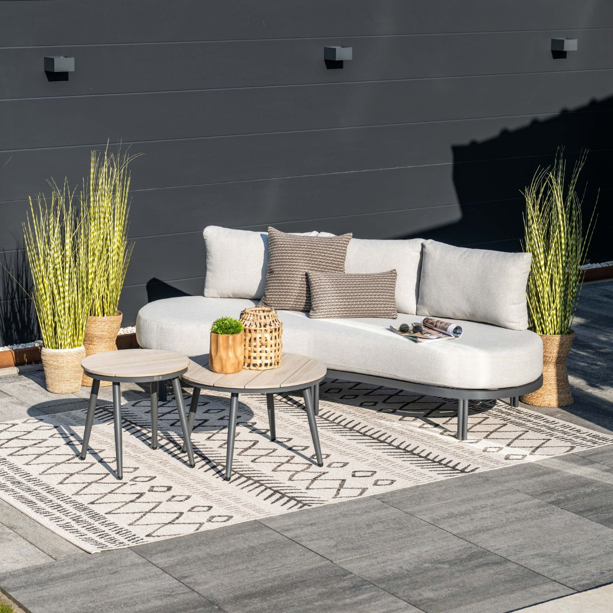 Moderne buitenbank met kussens, twee salontafels en planten tegen donkere muur.