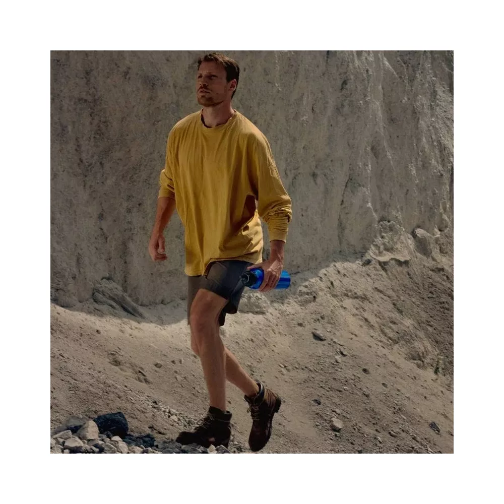 Mann in gelbem Pullover und Shorts geht in felsiger Landschaft mit blauer Wasserflasche.