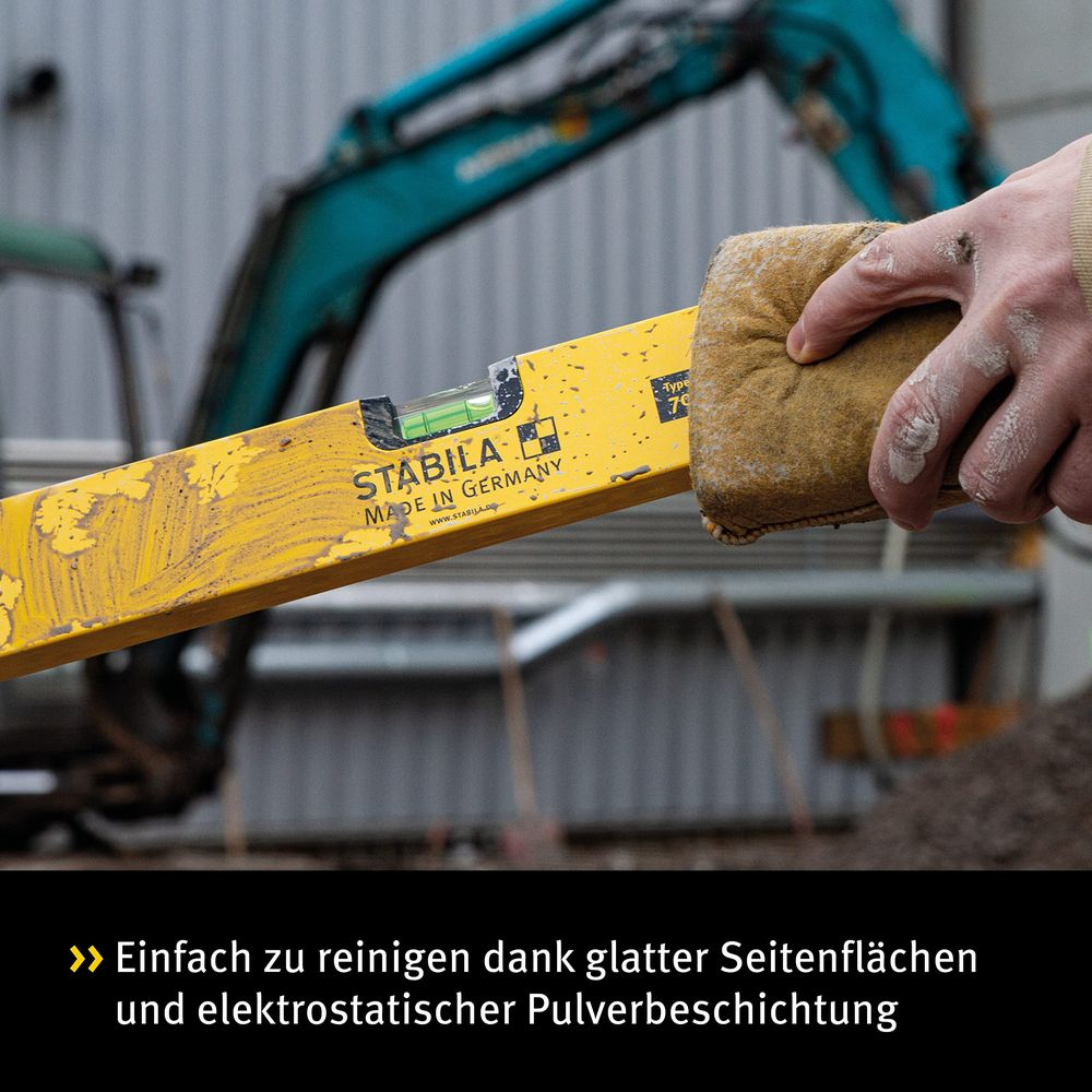 Eine Hand reinigt eine gelbe Wasserwaage mit einem braunen Tuch. Baustelle im Hintergrund.