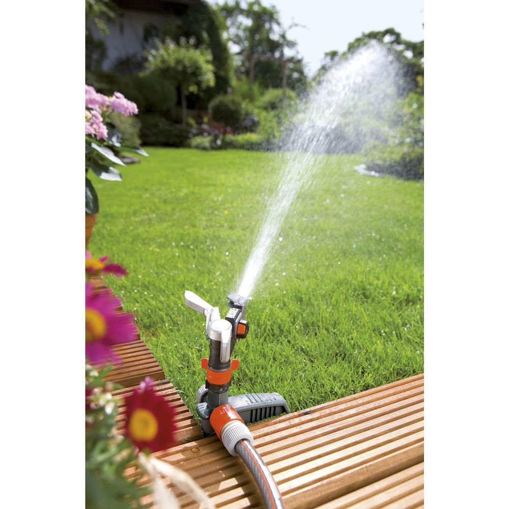 Ein Rasensprenger bewässert einen grünen Rasen mit einem Wasserstrahl. Holzdielen liegen davor.