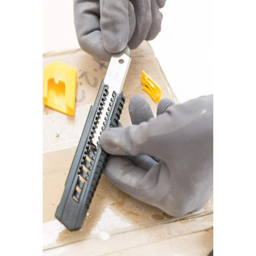 Graue Handschuhe halten ein Cuttermesser, gelbe Kappen liegen daneben.