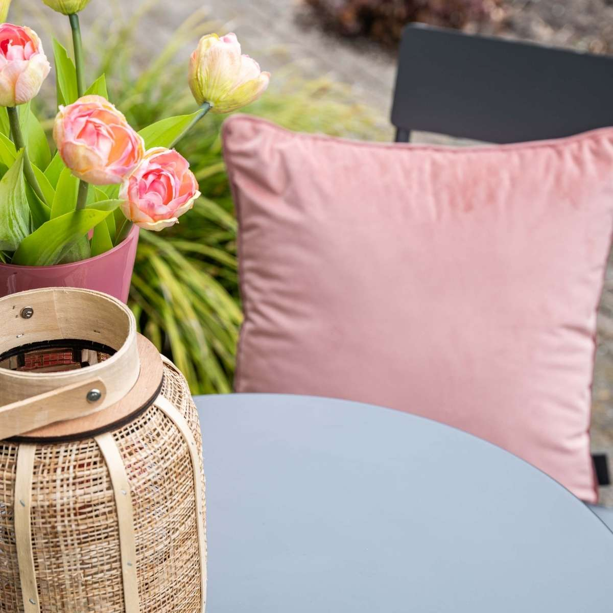 Een roze kussen op een stoel naast tulpen en een geweven lantaarn.