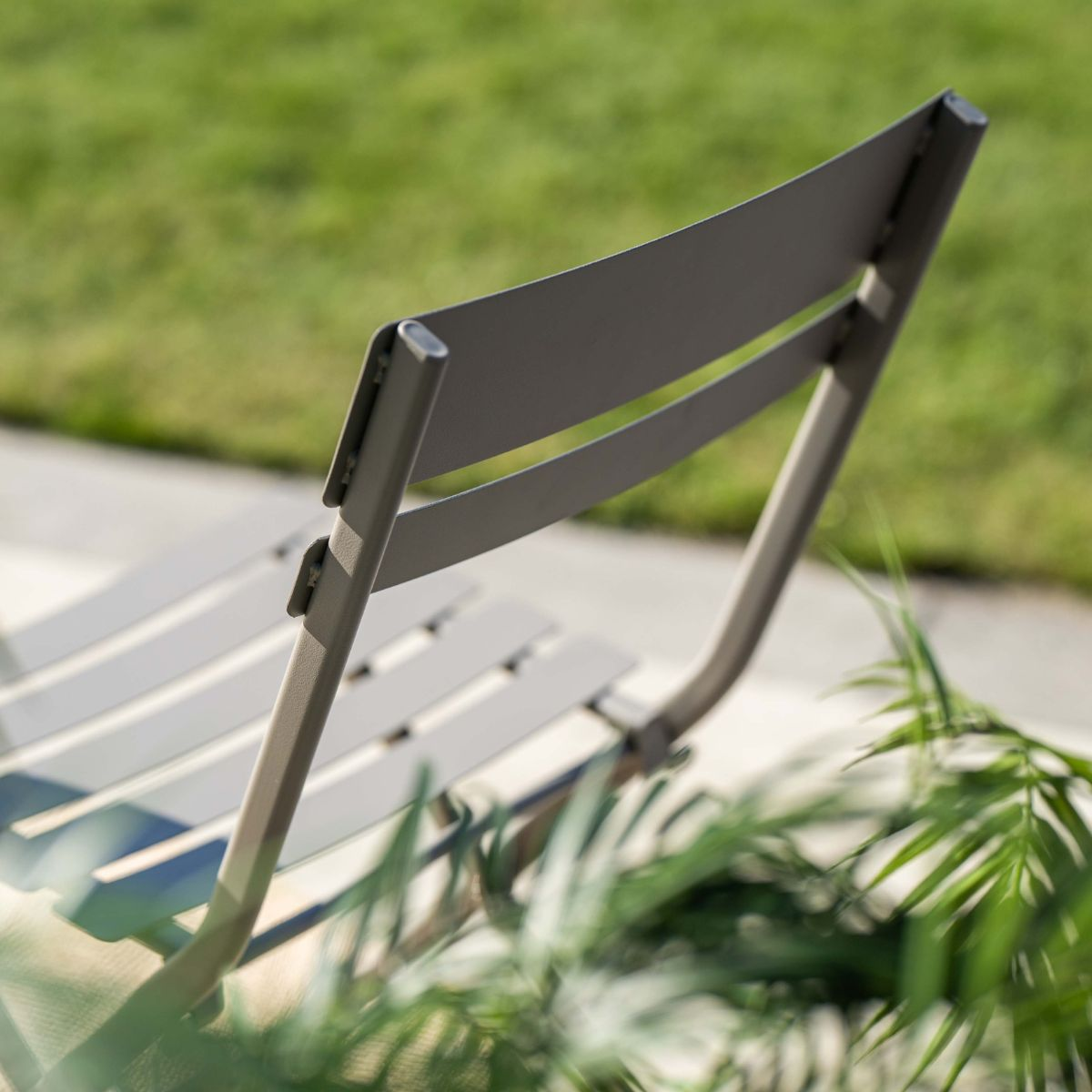 Grijze tuinstoel met lamellen op beton, met groen gras en planten op de achtergrond.