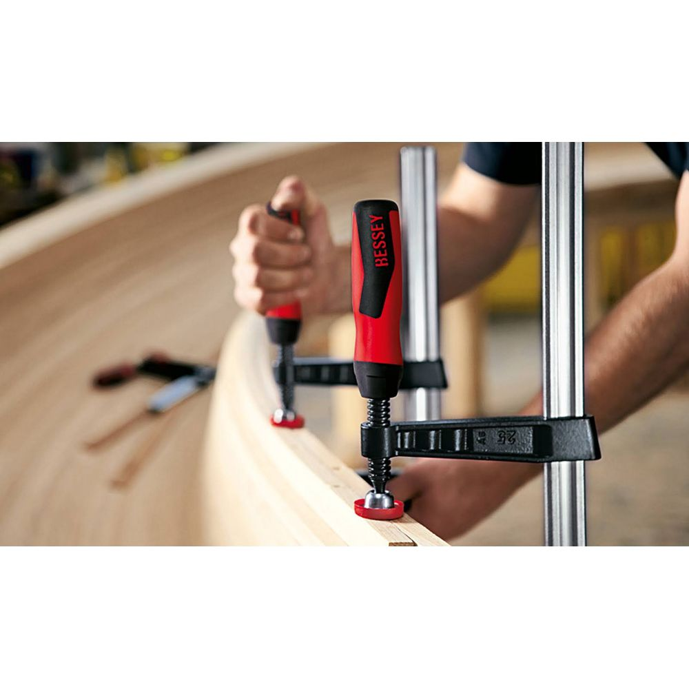 Eine Hand hält eine Bessey-Klemme mit rotem Griff, die ein Holzteil fixiert.
