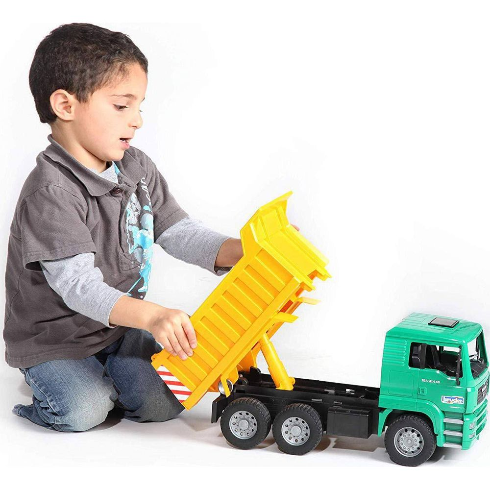 Chłopiec klęczy, bawiąc się żółtą skrzynią wywrotki przyczepioną do zielonej ciężarówki.