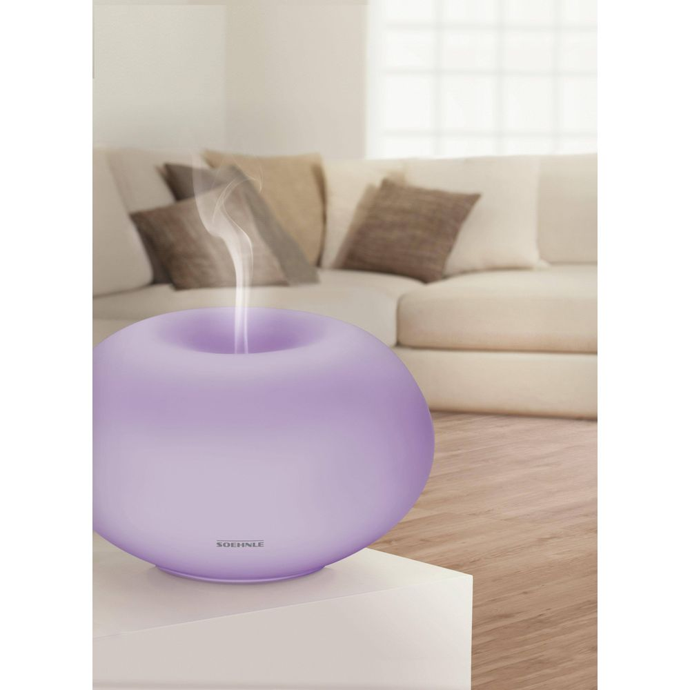 Ein lila Diffusor gibt Dampf ab, vor einem Sofa mit Kissen.