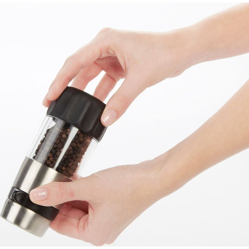 Dłonie trzymające przezroczysty szklano-metalowy młynek do pieprzu z czarnym pieprzem.