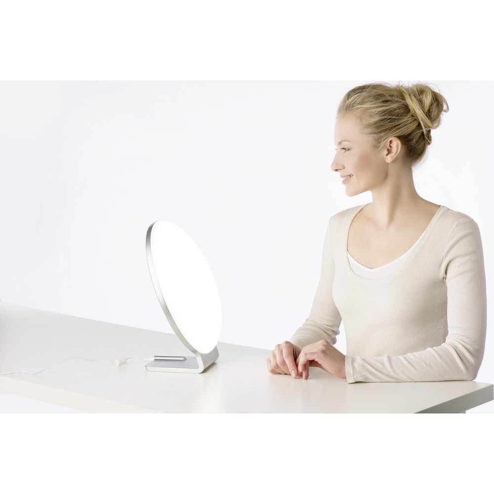 Eine Frau sitzt an einem Tisch und blickt auf eine Lichttherapielampe.