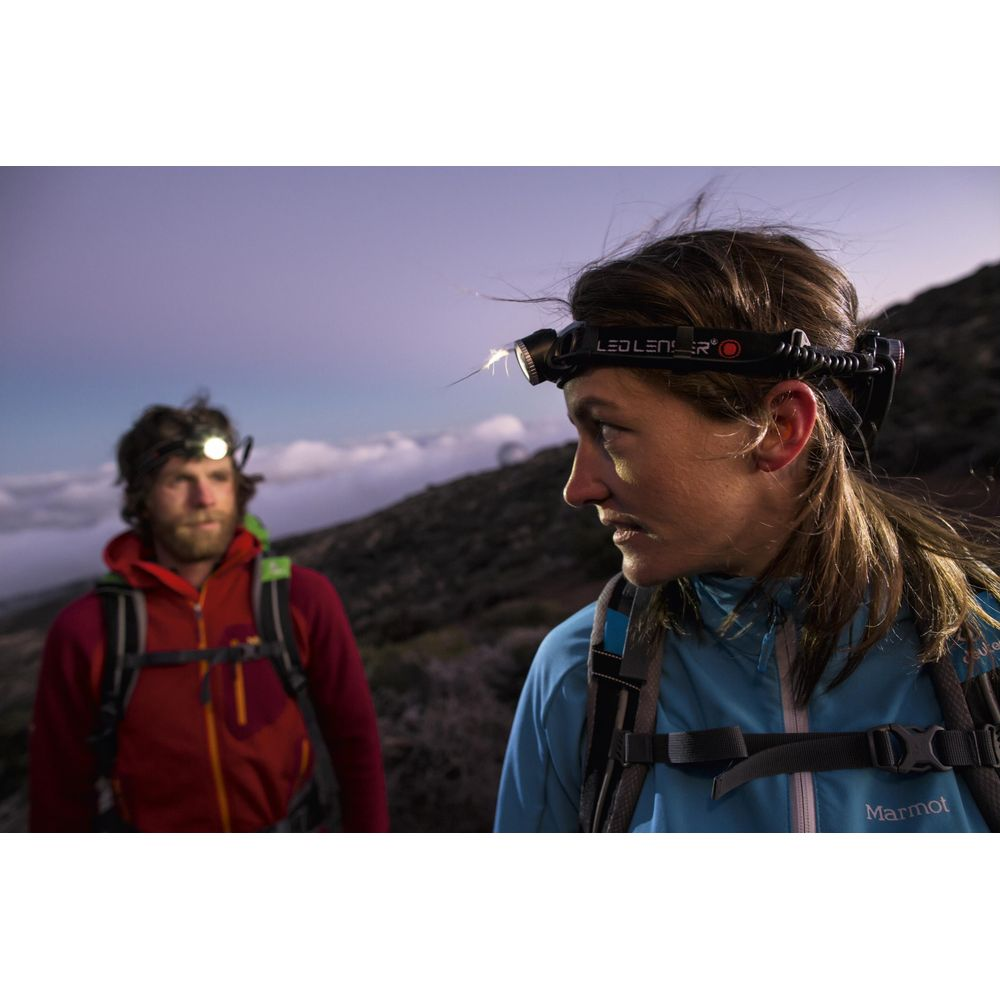 Deux randonneurs portent des lampes frontales sur un sentier de montagne au crépuscule. Femme en veste bleue, homme en rouge.