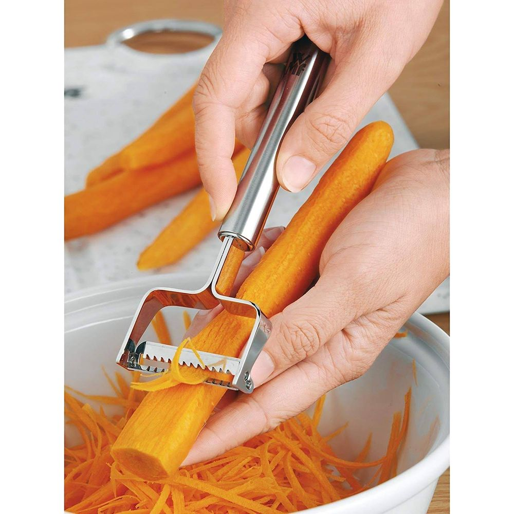 Des mains pelant une carotte avec un économe dans un bol de carottes râpées.