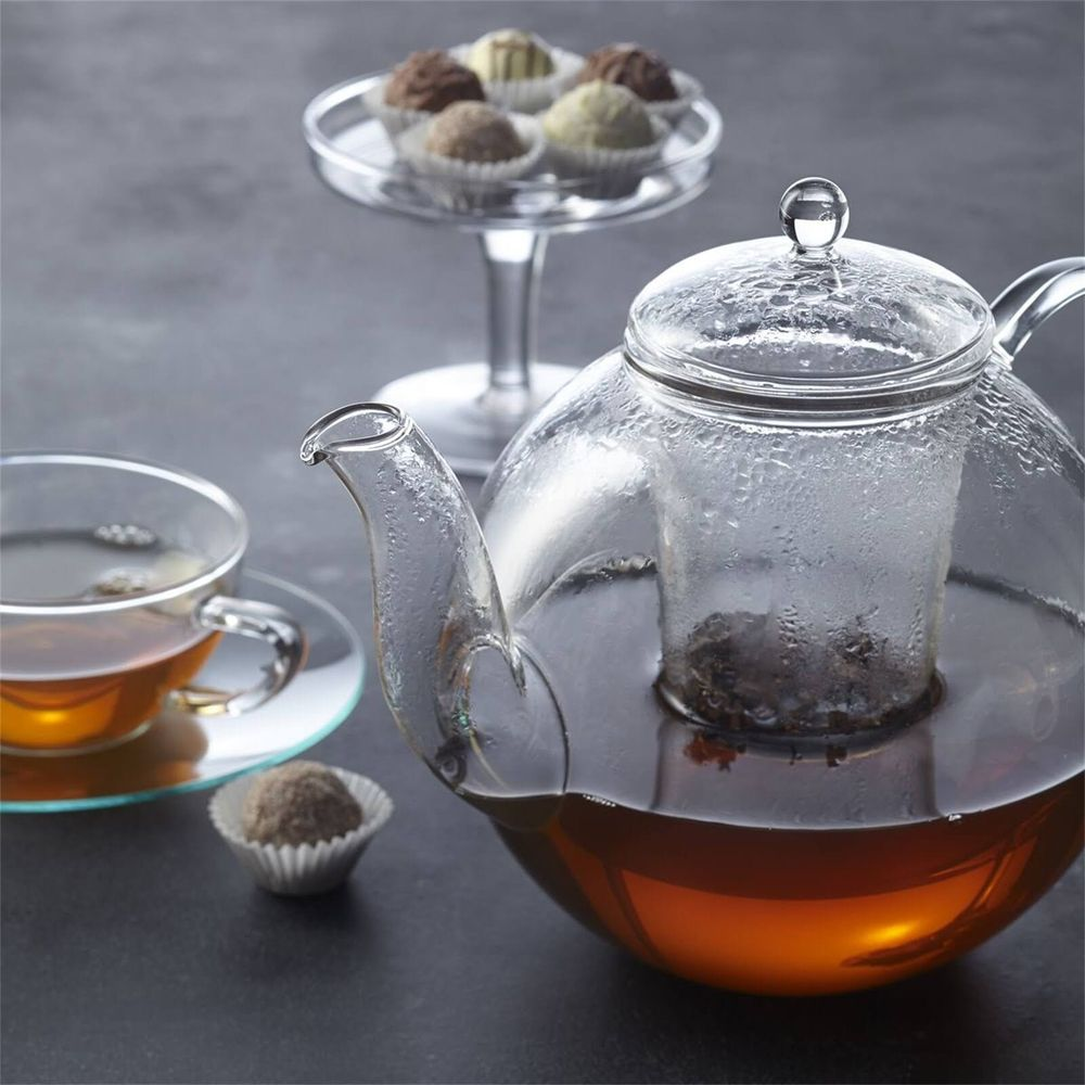 Ein Glas-Teekanne mit Tee und eine Tasse Tee daneben. Schokoladenpralinen im Hintergrund.